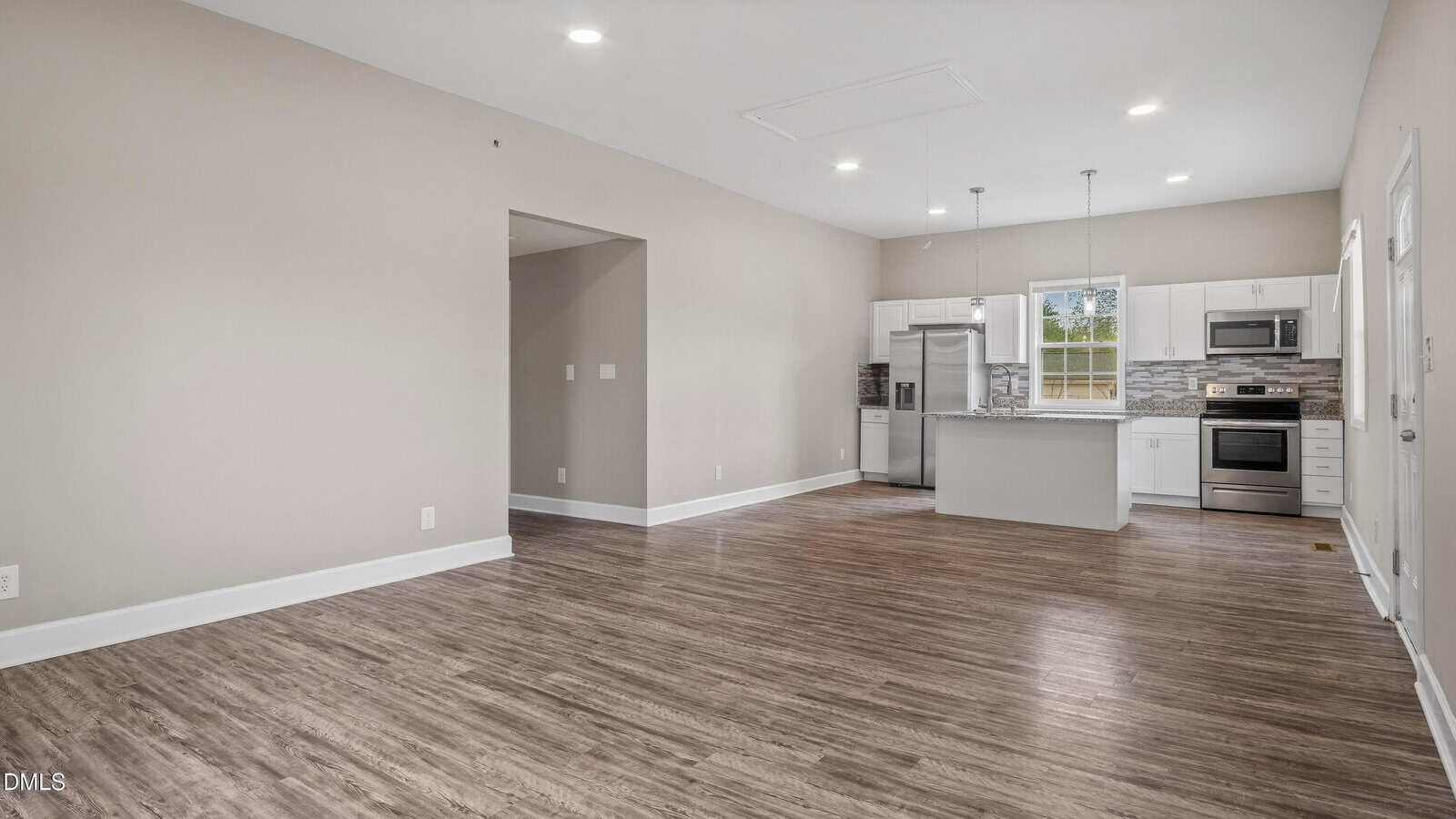 Open concept living area with wood floors, beige walls, and a modern kitchen featuring white cabinets, stainless steel appliances, a center island, and pendant lighting. Large windows provide natural light.