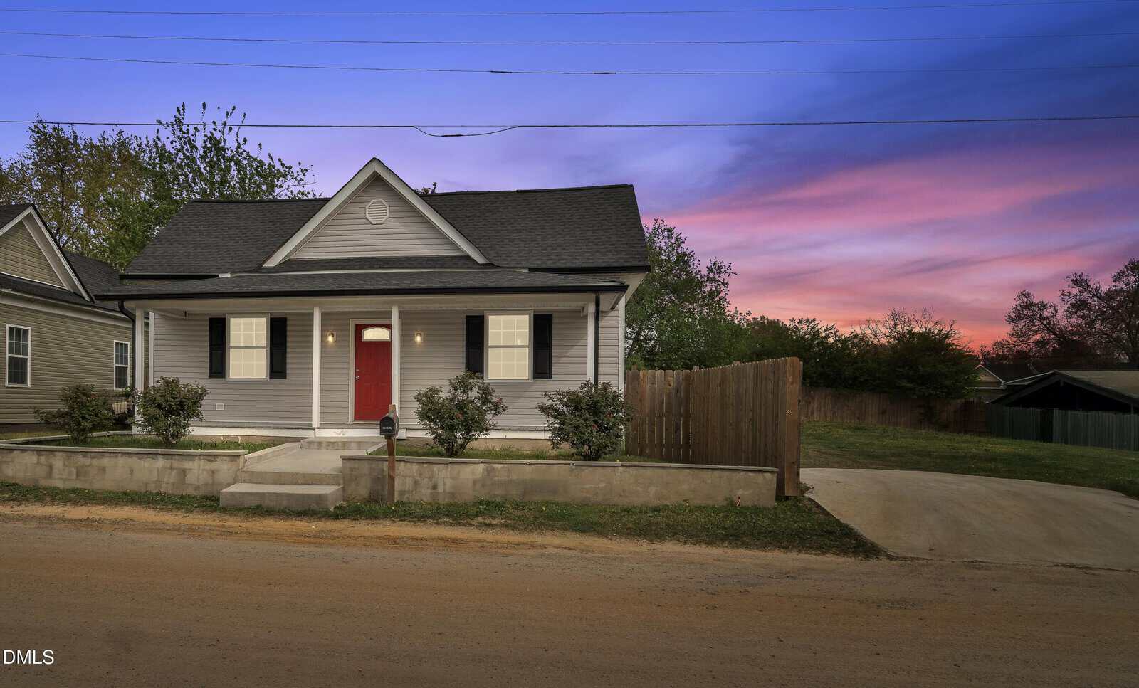 A small gray house at 1284 Railroad Road, Henderson, features a red front door, black shutters, and a covered porch. At dusk, enjoy the purple and pink sunset sky above the concrete walkway, shrubs, driveway, and wooden fence.