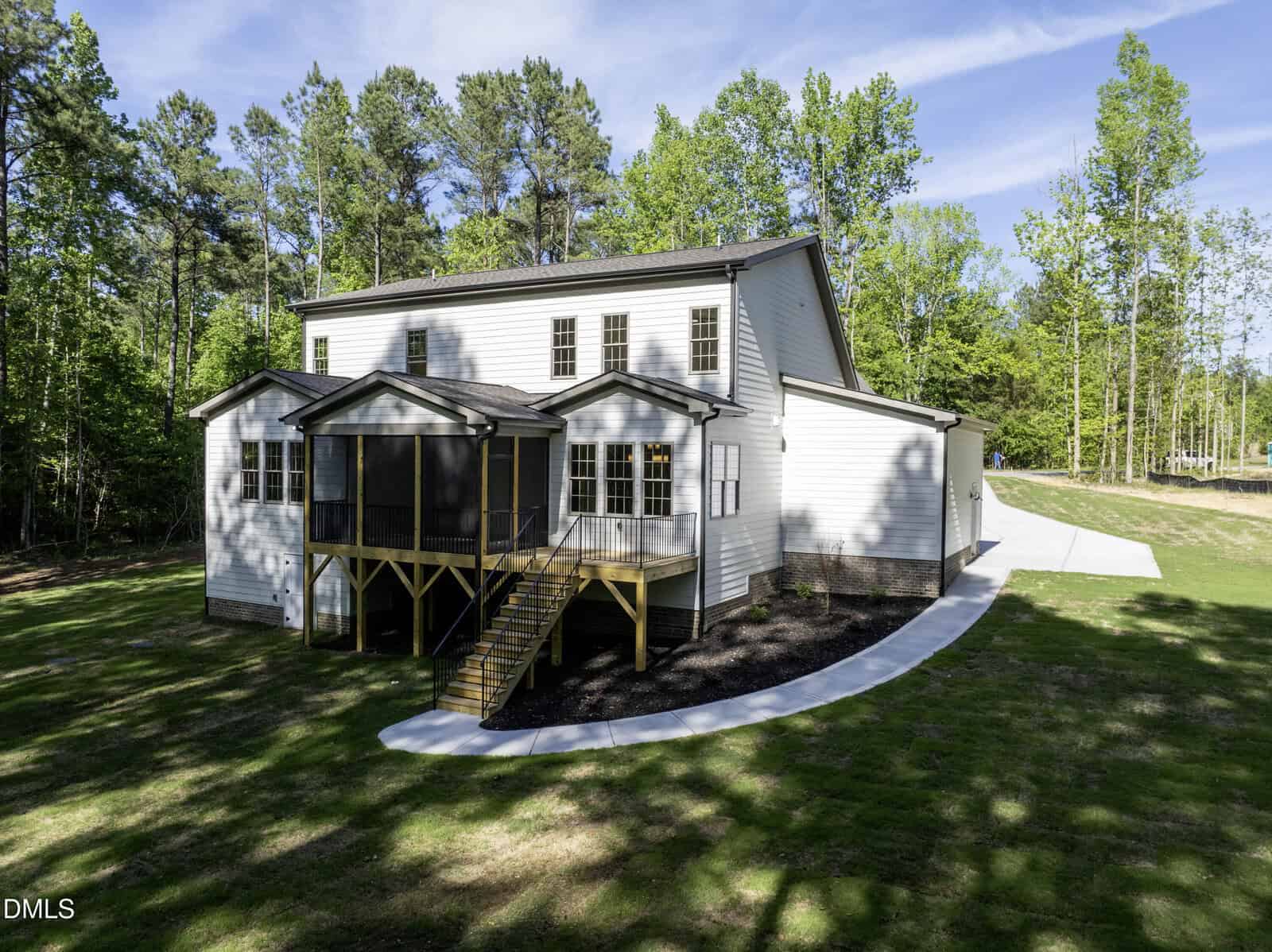 A two-story white house with a screened porch and wooden stairs, surrounded by a lawn and tall trees, with a concrete walkway curving around the yard on a sunny day.