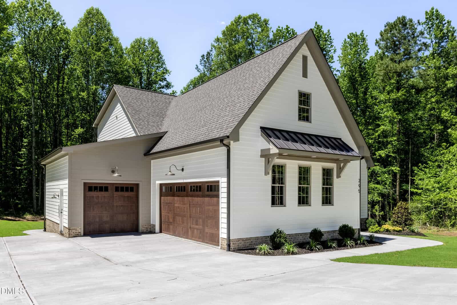 A modern detached garage with two wooden doors, white siding, gray roof, and small windows, surrounded by a concrete driveway and green trees on a sunny day.