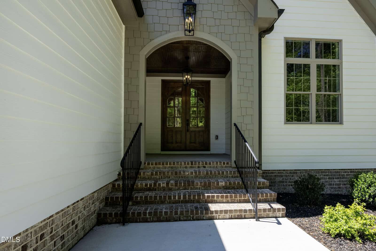 A front entrance of a house with brick steps, black metal railings, double wooden doors, hanging lantern lights, cream-colored siding, and a window to the right. Shrubs line the walkway.