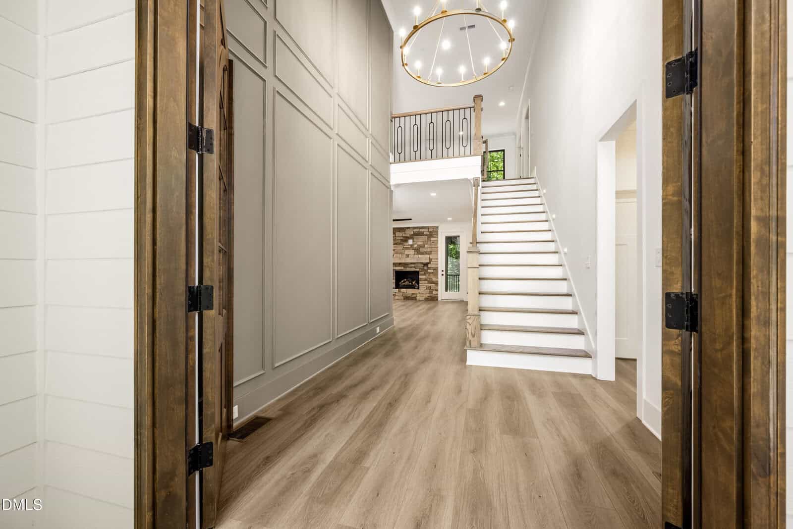 Spacious home entryway with wood floors, double doors, high ceilings, large chandelier, staircase with white risers and wooden railing, and a view into a living area with a stone fireplace.