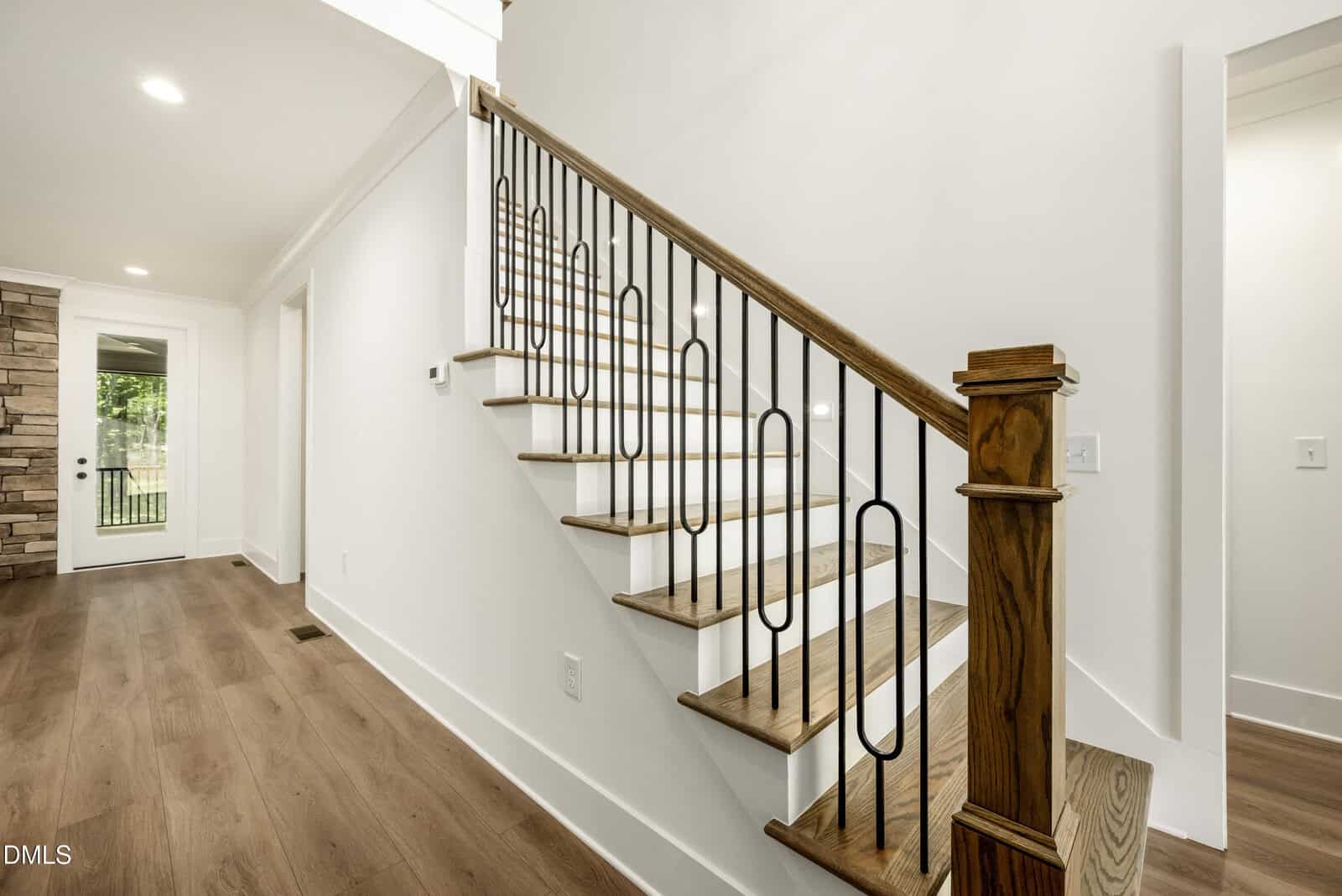 A wooden staircase with black metal balusters and a wooden handrail in a modern home; light wood flooring, white walls, and a doorway leading to a room with a stone accent wall are visible.