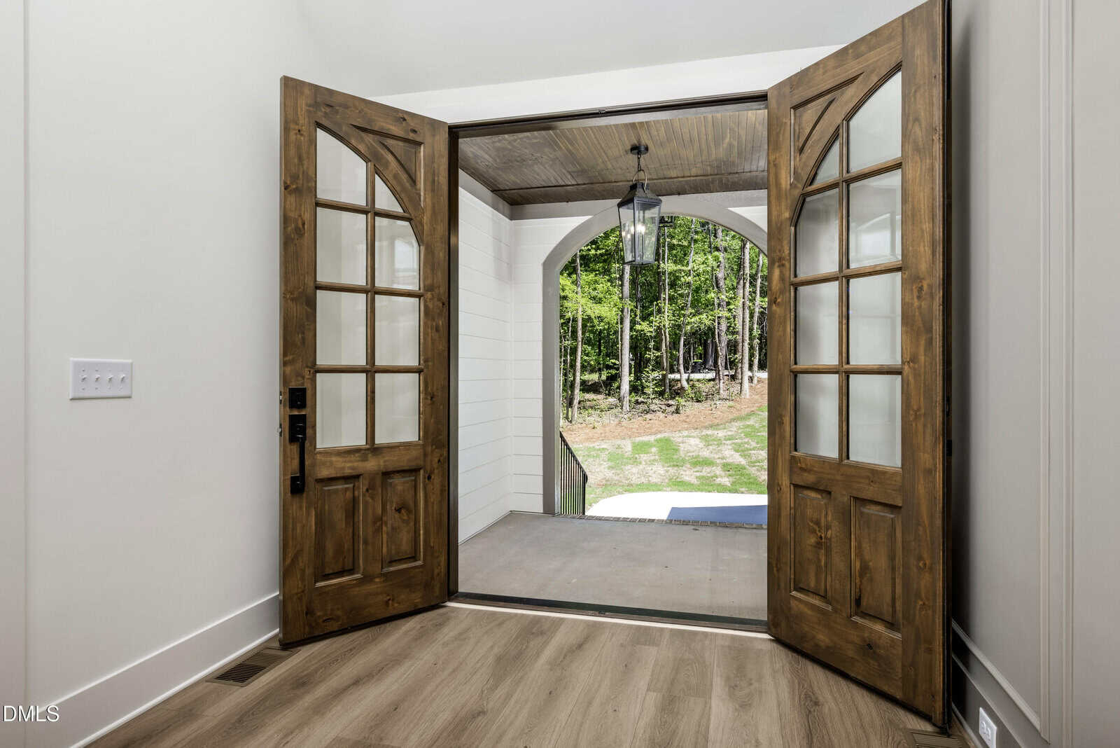 Double wooden doors with glass panels open to a covered porch. A hanging lantern is visible outside, with a view of a green yard and trees. The interior has light walls and wood flooring.