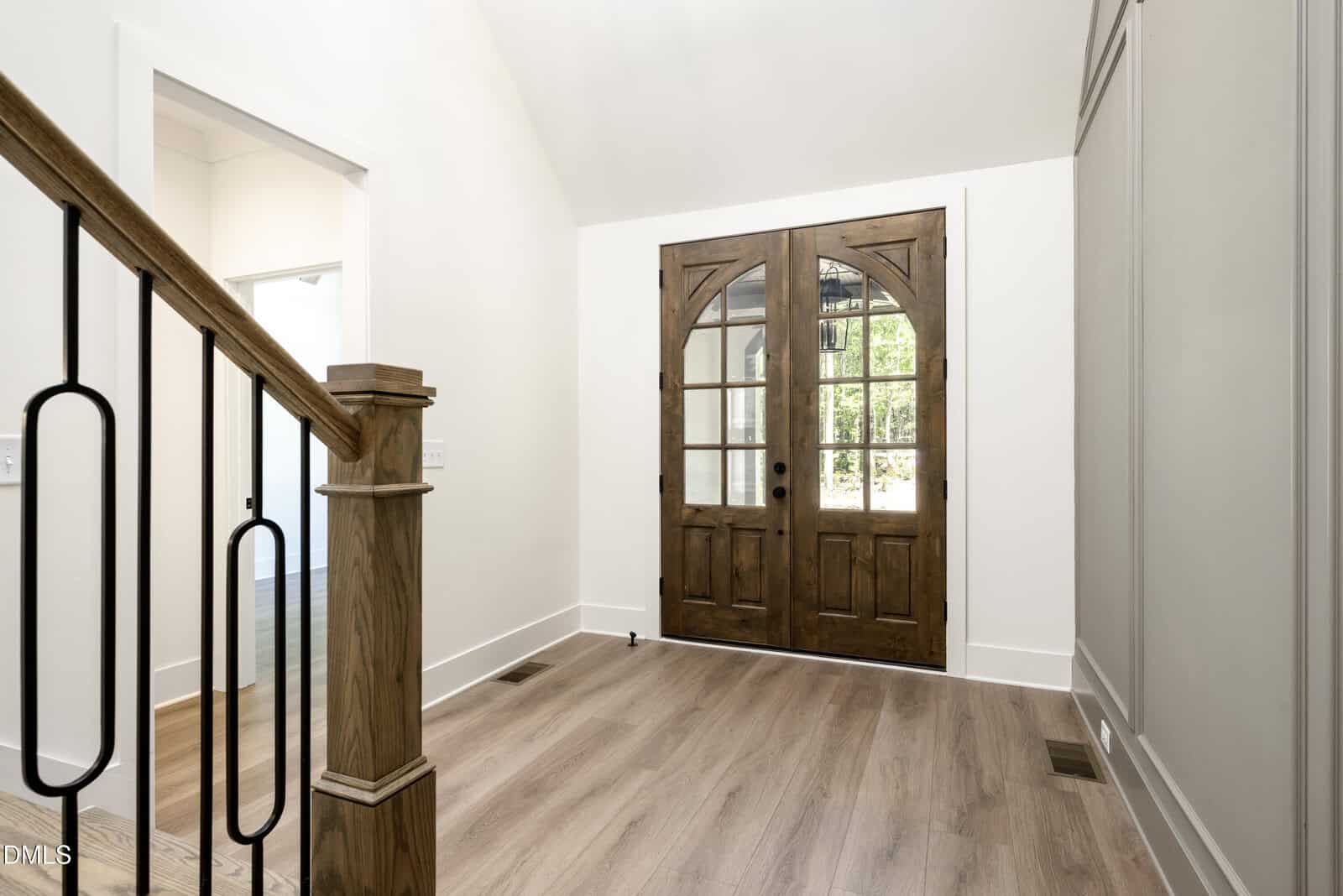 A bright entryway with light wood floors, white walls, a wooden staircase railing on the left, and double wooden front doors with glass panes. Natural light streams in through the doors' windows.
