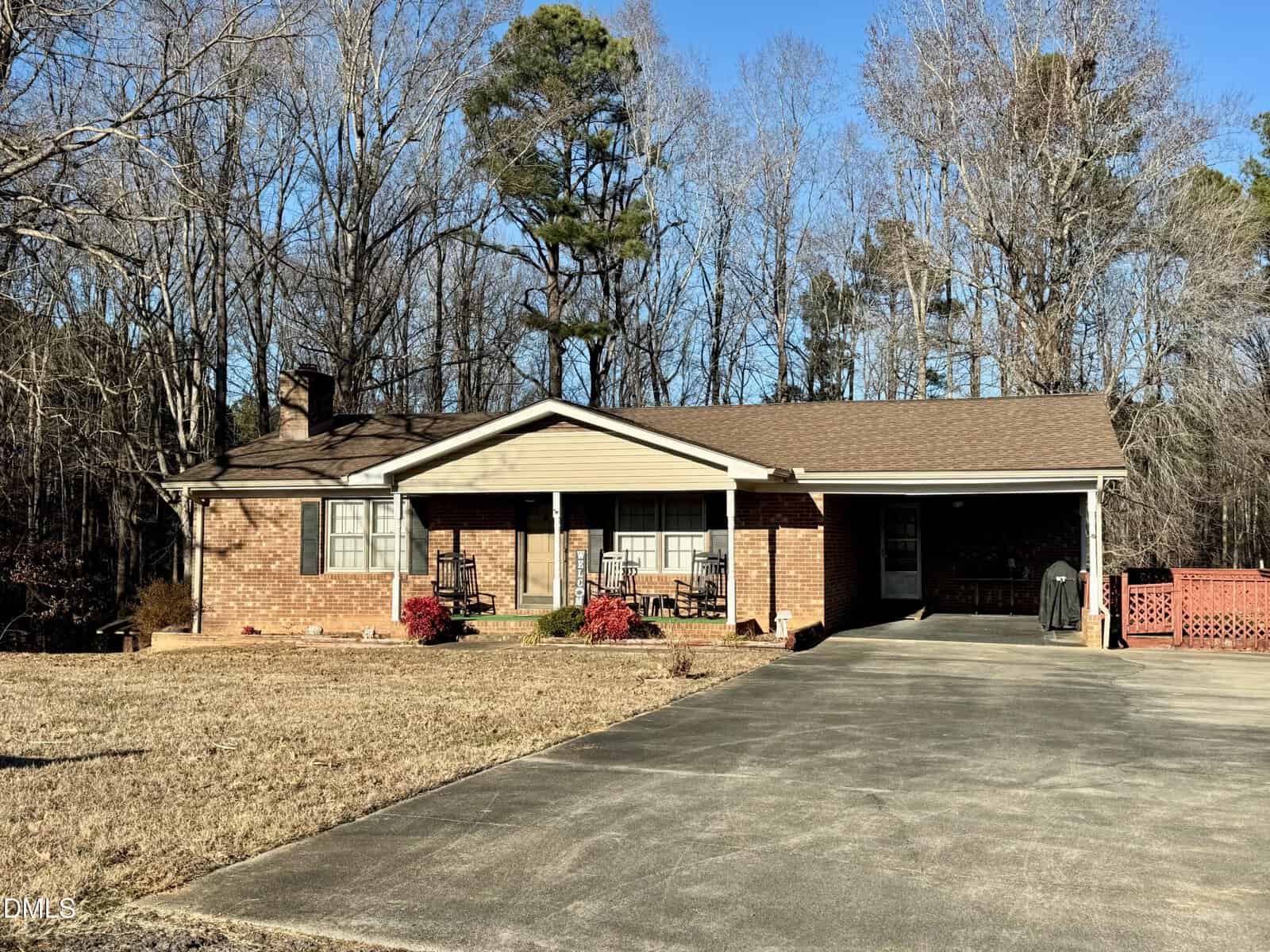 Single-story brick house with a front porch, rocking chairs, and attached carport. The yard is grassy and bordered by leafless trees under a clear blue sky.