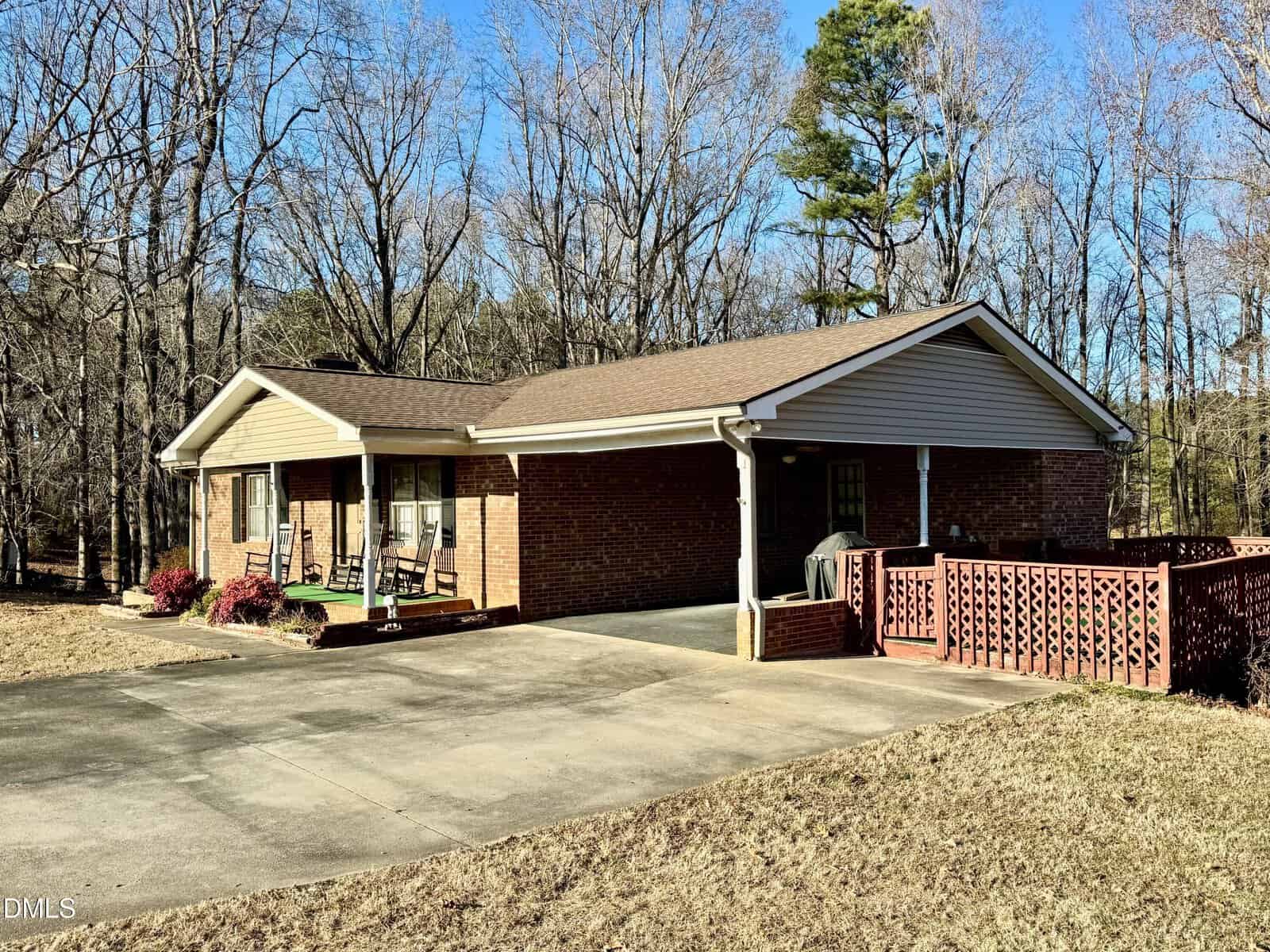 A single-story brick house with a covered porch, rocking chairs, a carport, and a red wooden railing. The yard is dry and surrounded by leafless trees on a clear, sunny day.