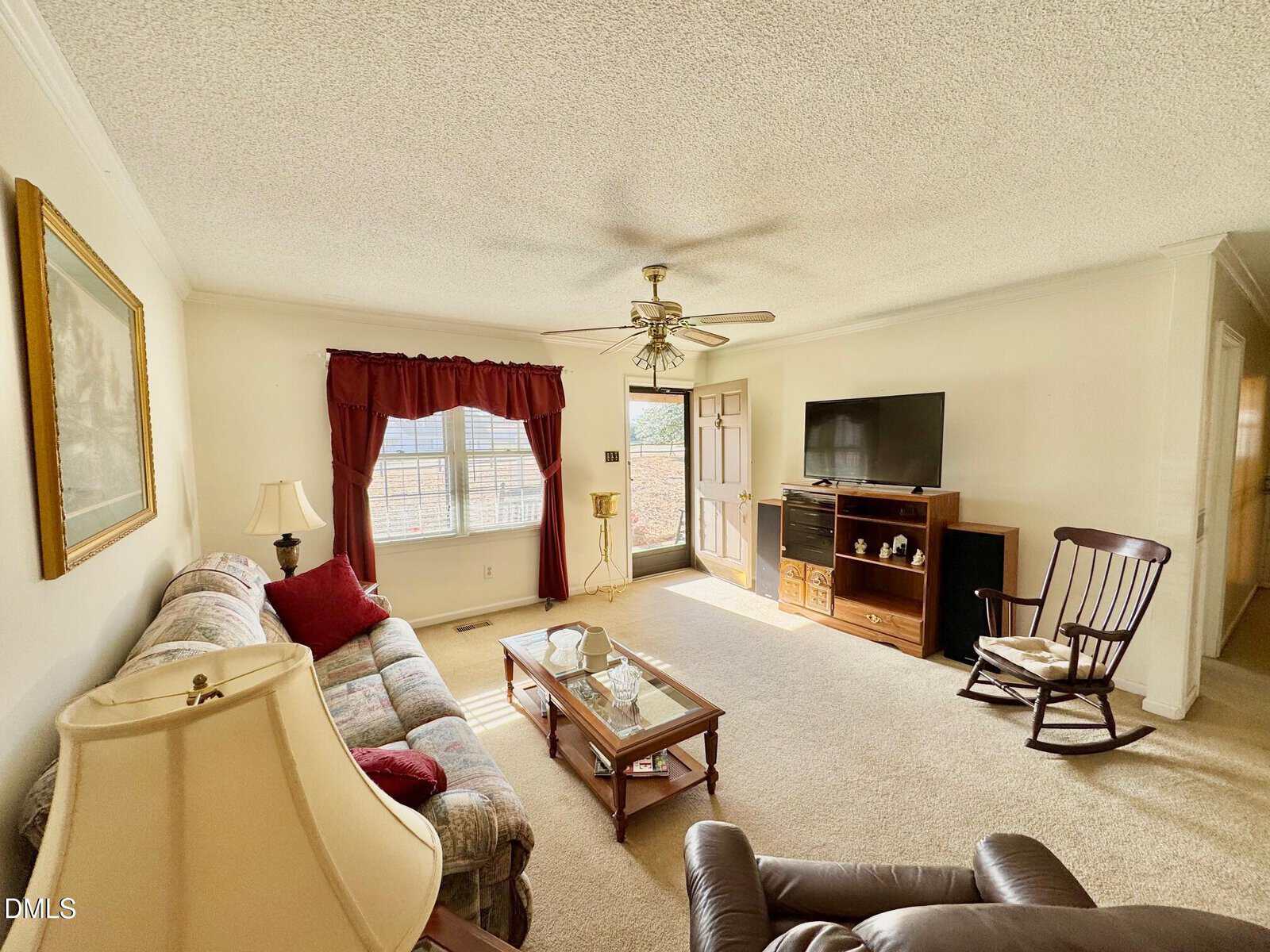 Bright living room with beige carpet, floral sofa, glass coffee table, rocking chair, wooden TV stand with TV, and a large window with red curtains near the front door. Ceiling fan overhead, lamp, and wall art visible.