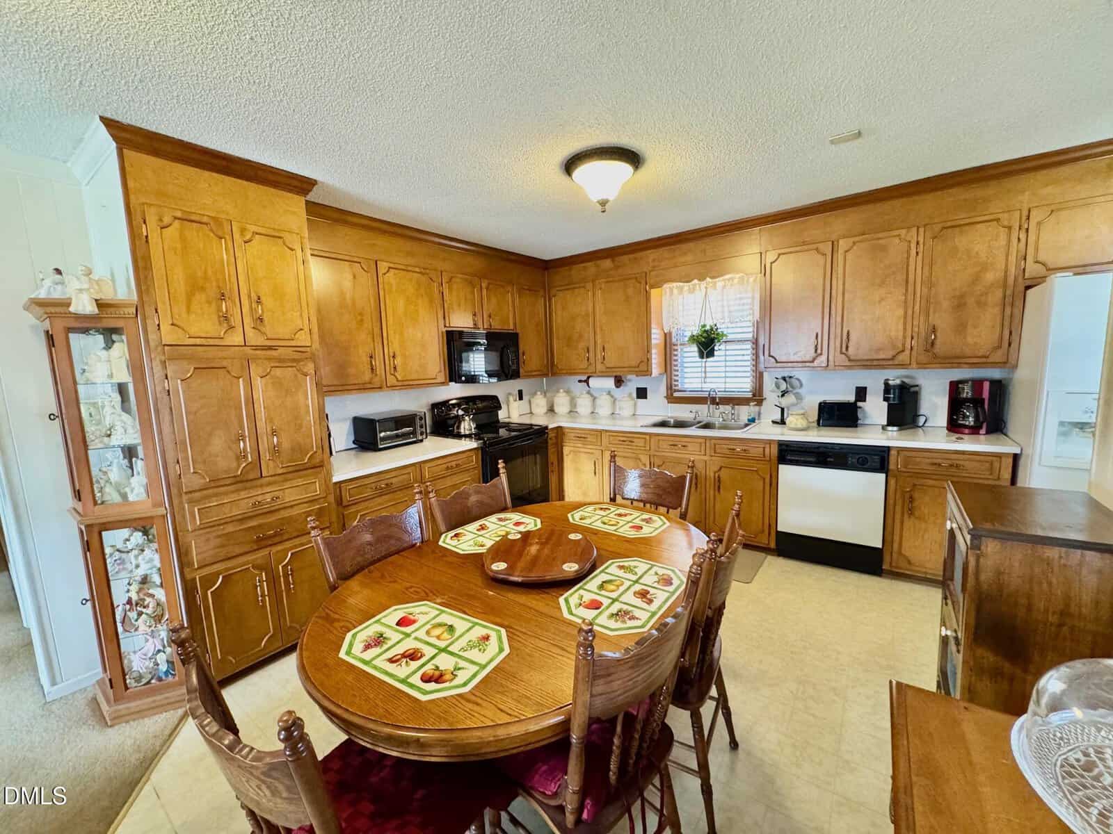 A cozy kitchen with wooden cabinets, a round dining table set for four, various appliances, and sunlight streaming through a window with blinds above the sink.