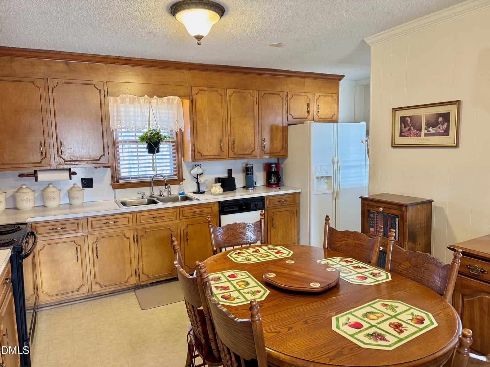 A cozy kitchen with wooden cabinets, a round dining table set with floral placemats, and a white refrigerator. A window with a valance is above the sink, and a framed picture hangs on the cream-colored wall.