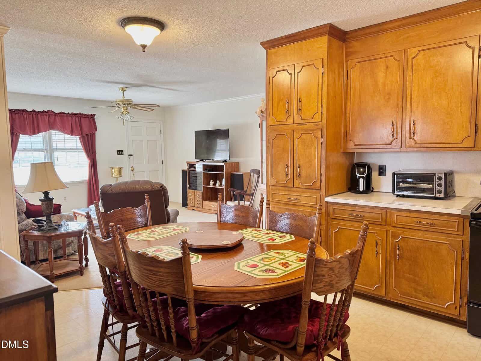 A cozy kitchen and dining area with a wooden table and six chairs, wooden cabinets, a coffee maker, toaster oven, and a view into a living room with a recliner, TV, and red curtains on the window.