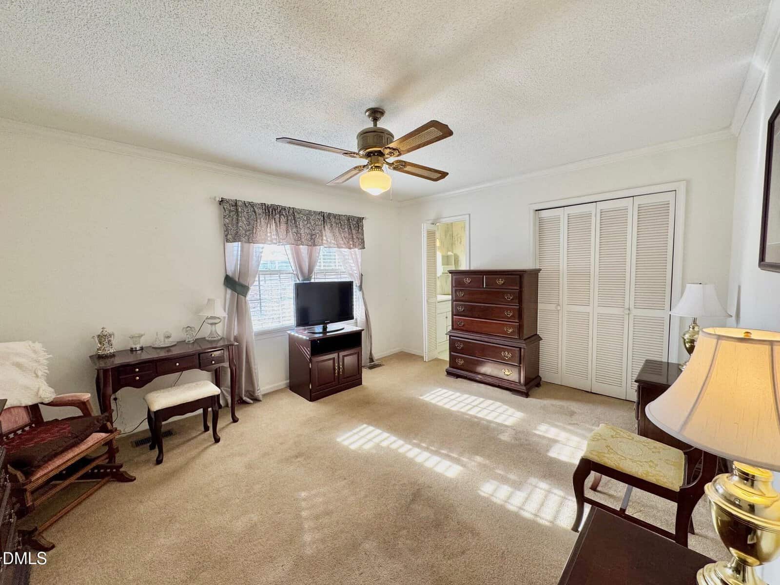 Bedroom with beige carpet, wooden furniture, TV on a stand, window with curtains, ceiling fan, chest of drawers, and a closet with louvered doors. Sunlight streams in through the window onto the floor.