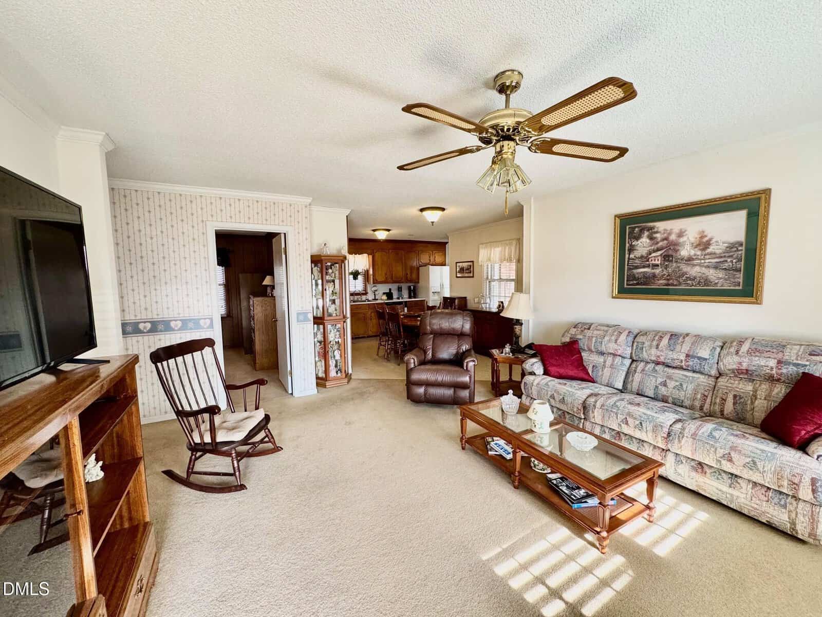 A cozy living room with a patterned sofa, glass coffee table, armchair, TV, rocking chair, ceiling fan, and a kitchen area in the background. Sunlight streams in through unseen windows.
