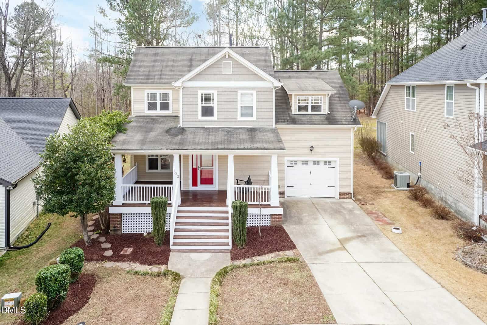 A two-story beige house with white trim at 102 Kipling Drive, Oxford, features a front porch, red door, attached single-car garage, neat lawn, driveway, and tall trees in the background among neighboring homes.