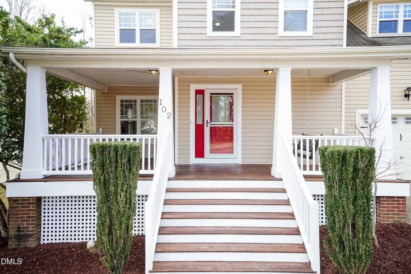 A front view of a beige house with white trim, a large covered porch, white railings, wooden steps, and a red front door. Two neatly trimmed shrubs frame the steps.