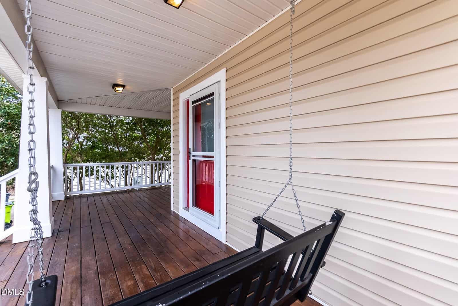 A wooden porch with beige siding, a black porch swing hanging from chains, a red door with a glass storm door, white railings, and a ceiling with recessed lights. Trees are visible in the background.