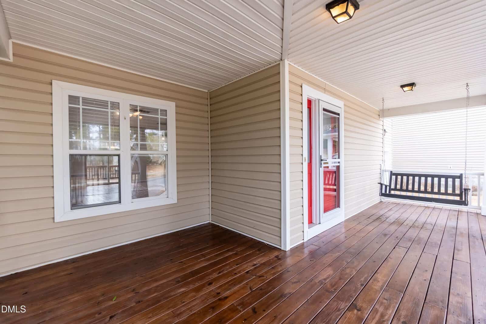 Covered front porch with beige siding, white trim, a red front door, two windows, wooden floor, ceiling lights, and a black porch swing.