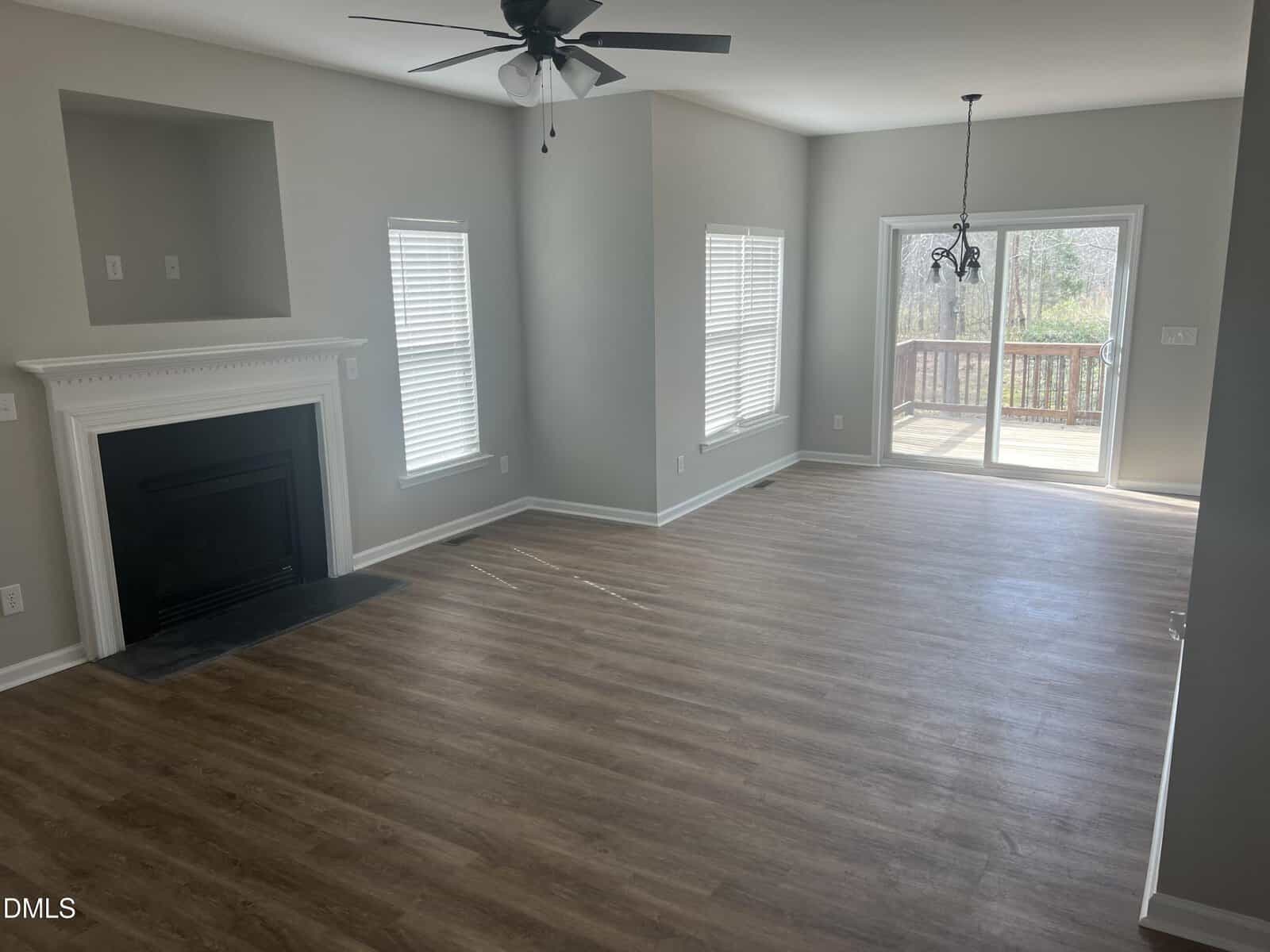 Bright, empty living room with wood floors, white fireplace, ceiling fan, two windows, and glass doors leading to a deck. Walls are light gray, and sunlight streams through the windows and doors.