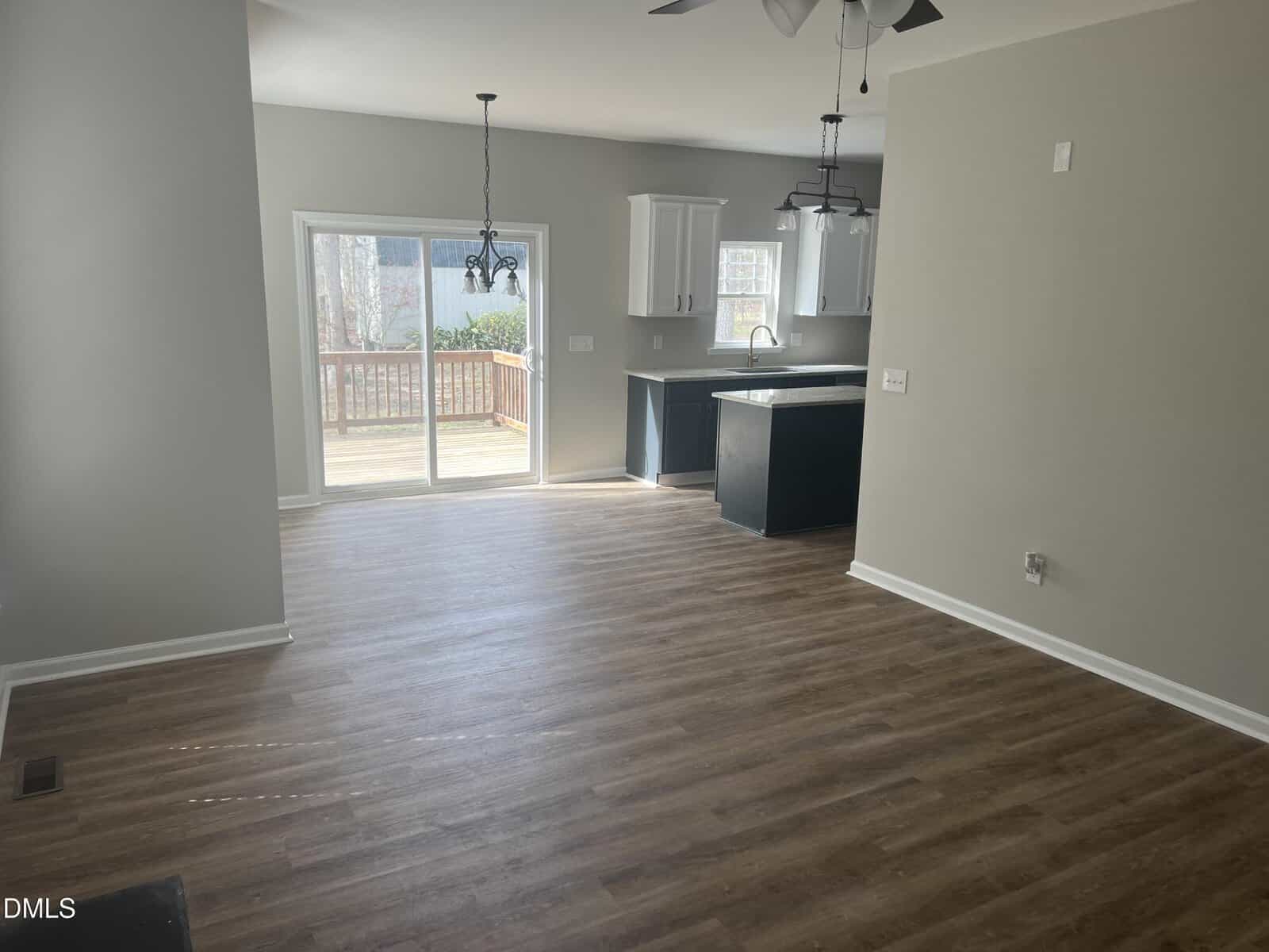 Empty room with wood flooring, beige walls, and an open layout leading to a kitchen with white cabinets and a dark island. Glass sliding doors open to a wooden deck outside. Ceiling fans and pendant lights are visible.