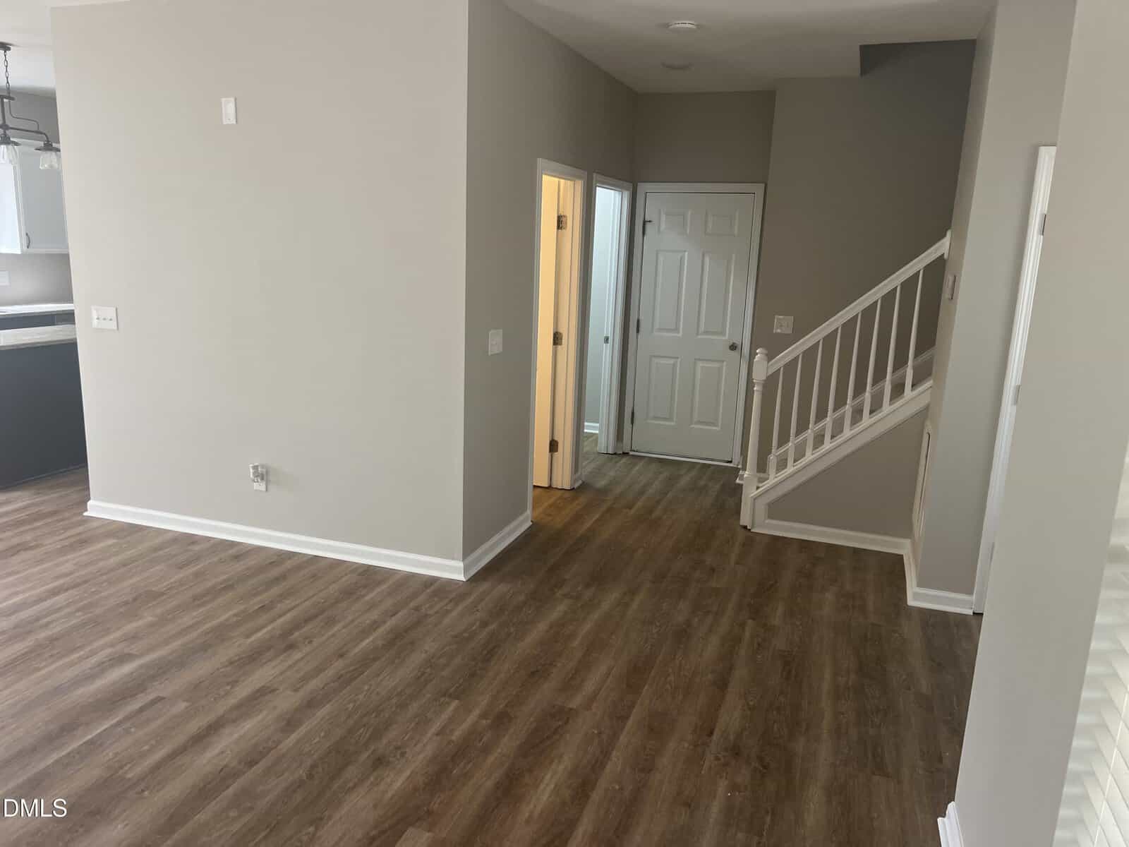 An empty room with wood floors, light beige walls, a staircase with white railing, a closed door, and an open doorway. The kitchen area is partially visible on the left.