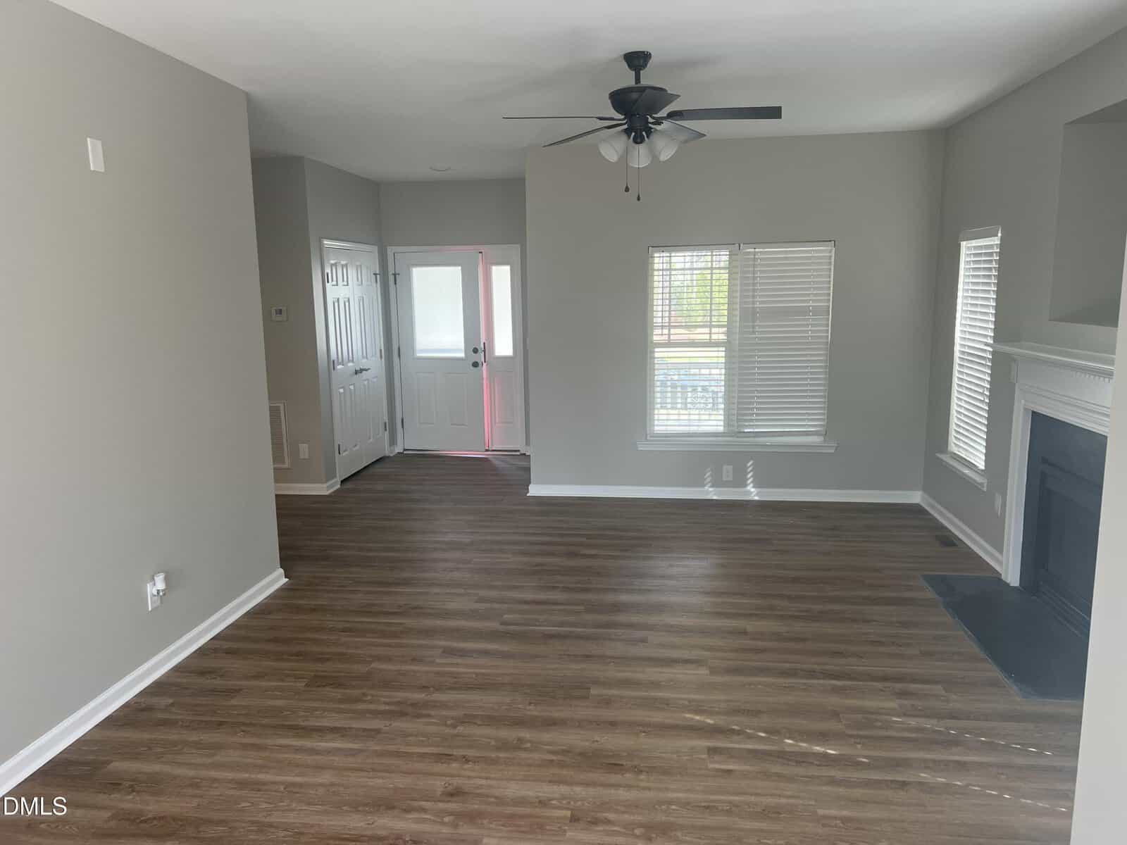 Empty living room with light gray walls, wood-look flooring, white trim, a ceiling fan, a fireplace on the right, and windows letting in natural light near a white front door.