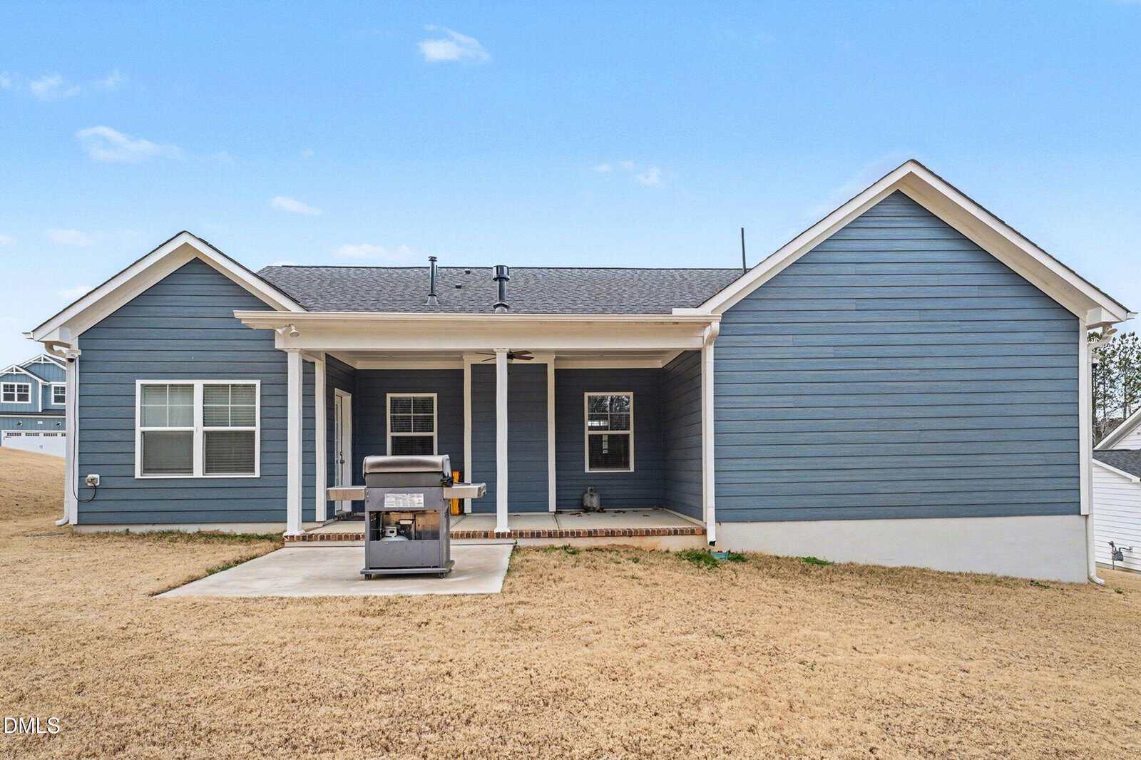 A single-story house with blue siding, white trim, and a covered back patio. A grill is placed on a small concrete pad in the backyard, which has dry, brown grass. The sky is clear with a few clouds.