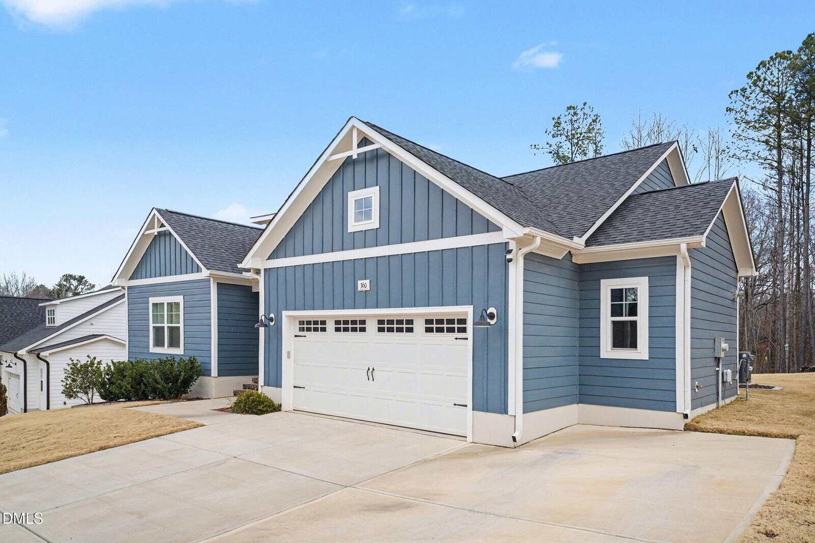A modern blue house with white trim, a double garage, and a wide driveway on a neatly maintained lawn, set against a clear blue sky and surrounded by trees.