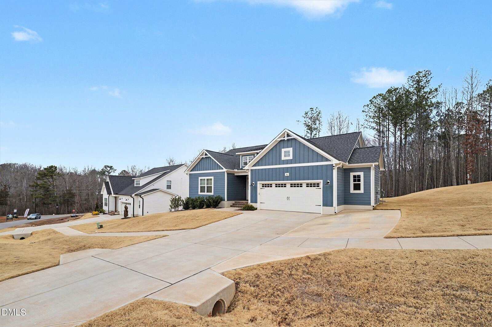 A modern blue house with white trim and a three-car garage sits on a large corner lot with a sloped, yellow-brown lawn, surrounded by trees and a clear sky in the background.