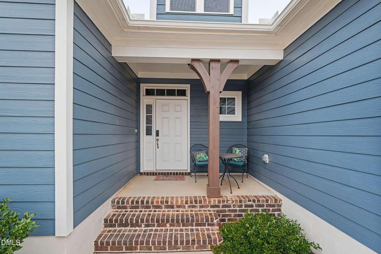 A small covered porch with brick steps leads to a white front door. The house exterior is blue with white trim. To the right of the door, there is a small round table with two chairs and cushions.