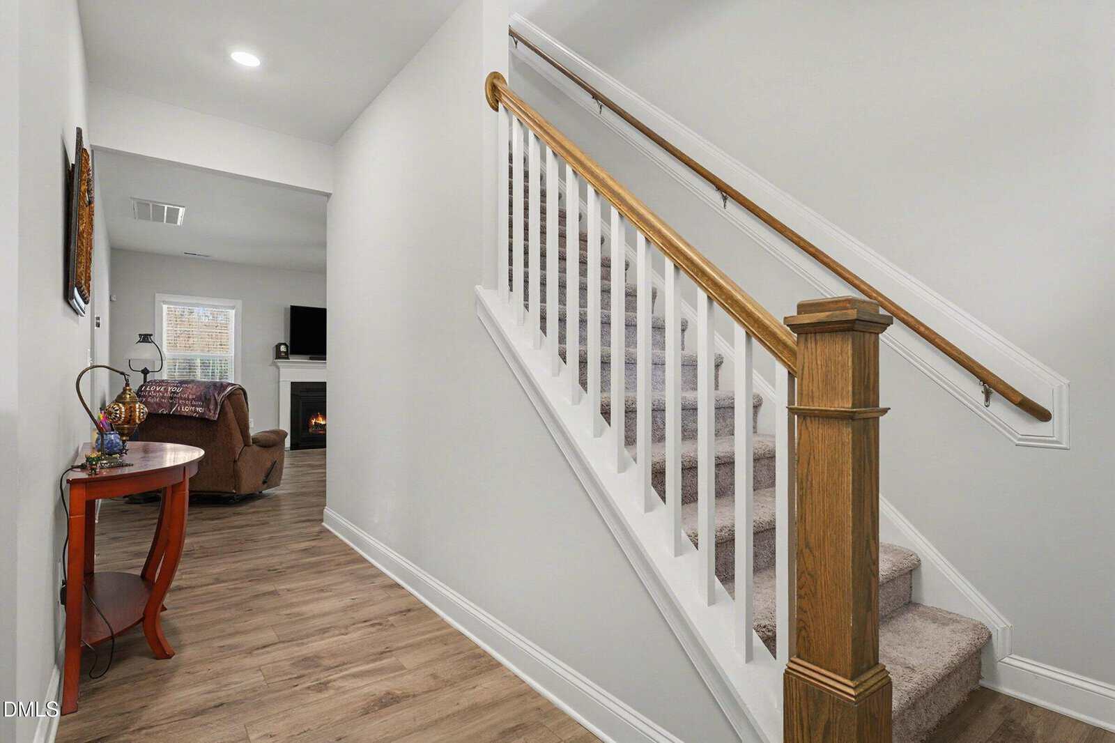 A carpeted staircase with a wooden handrail leads up from a hallway with light-colored walls and wood flooring. A small table with decor sits on the left, and a living room with a fireplace is visible in the background.
