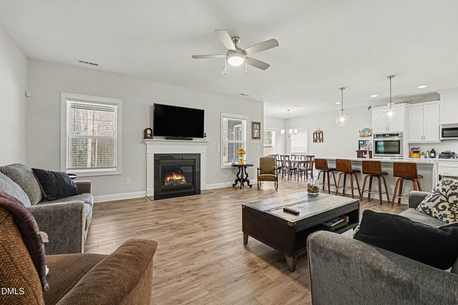 Modern open-concept living room and kitchen with wood flooring, gray sofas, a fireplace under a wall-mounted TV, large windows, ceiling fans, and a kitchen island with bar stools and pendant lights.