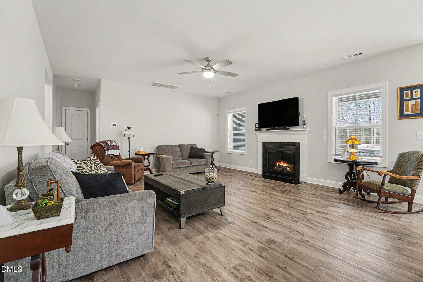 A cozy living room with gray sofas, a coffee table, an armchair, and a fireplace beneath a wall-mounted TV. The room has wood flooring, a ceiling fan, and windows letting in natural light.