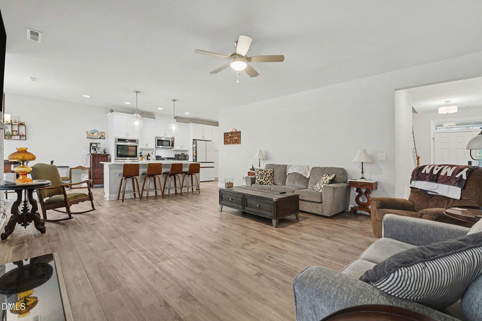 Open-layout living room and kitchen with wood floors, neutral furniture, ceiling fan, bar stools at kitchen island, and various lamps and decor. White walls and natural light create a bright, spacious atmosphere.