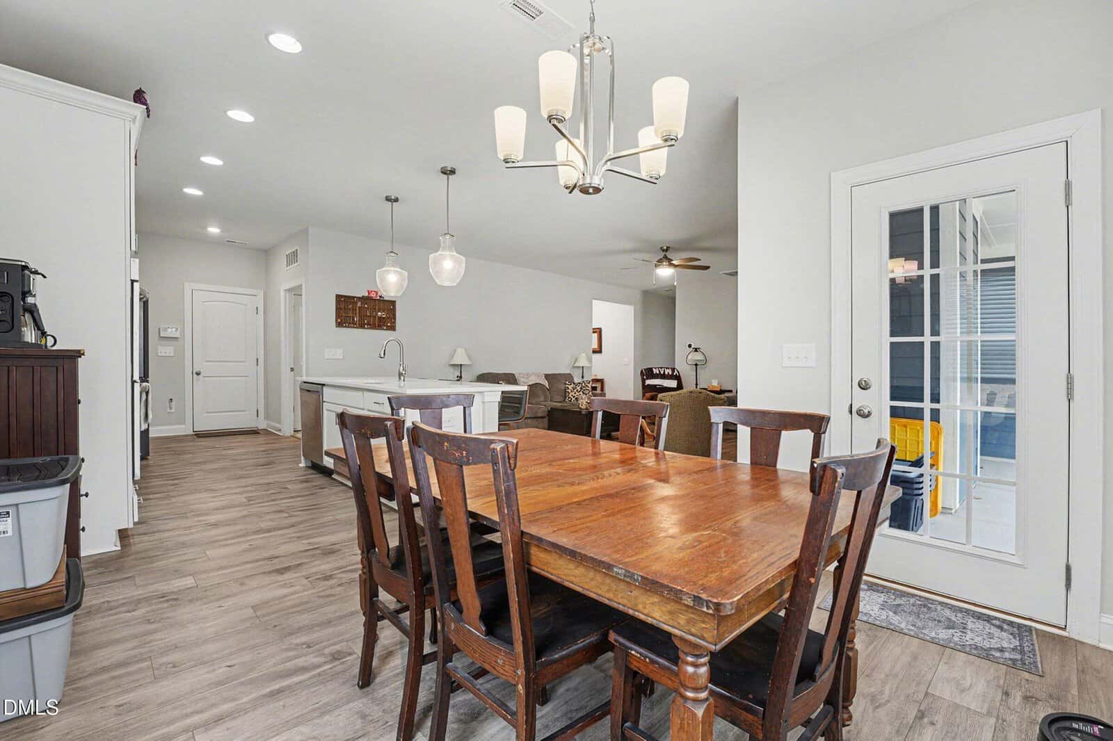 Dining area with a wooden table and six chairs, modern light fixture above, open to a kitchen with white cabinets, island, and pendant lights. Living room visible in the background, and a glass door leads outside.