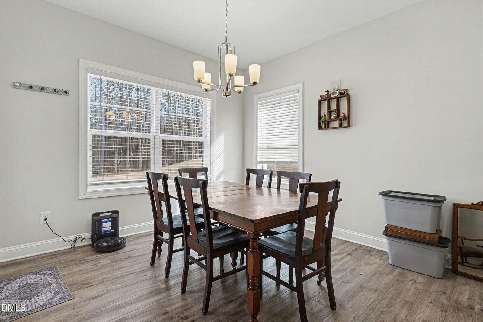 Bright dining room with six wooden chairs around a rectangular table, chandelier above, large windows with blinds, light gray walls, and minimal wall decorations. Plastic bins and a small device are in the corner.