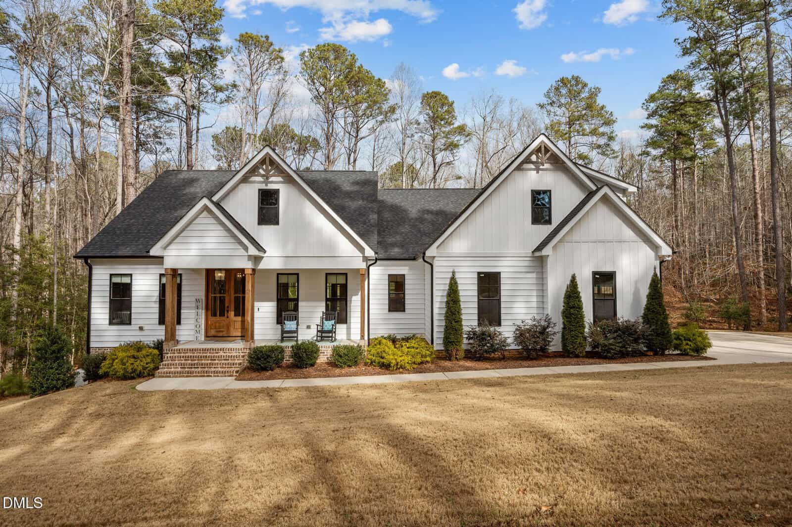 White modern farmhouse with black accents, front porch with wooden columns, manicured landscaping, and a large front yard with brown grass, surrounded by tall pine trees under a blue sky.
