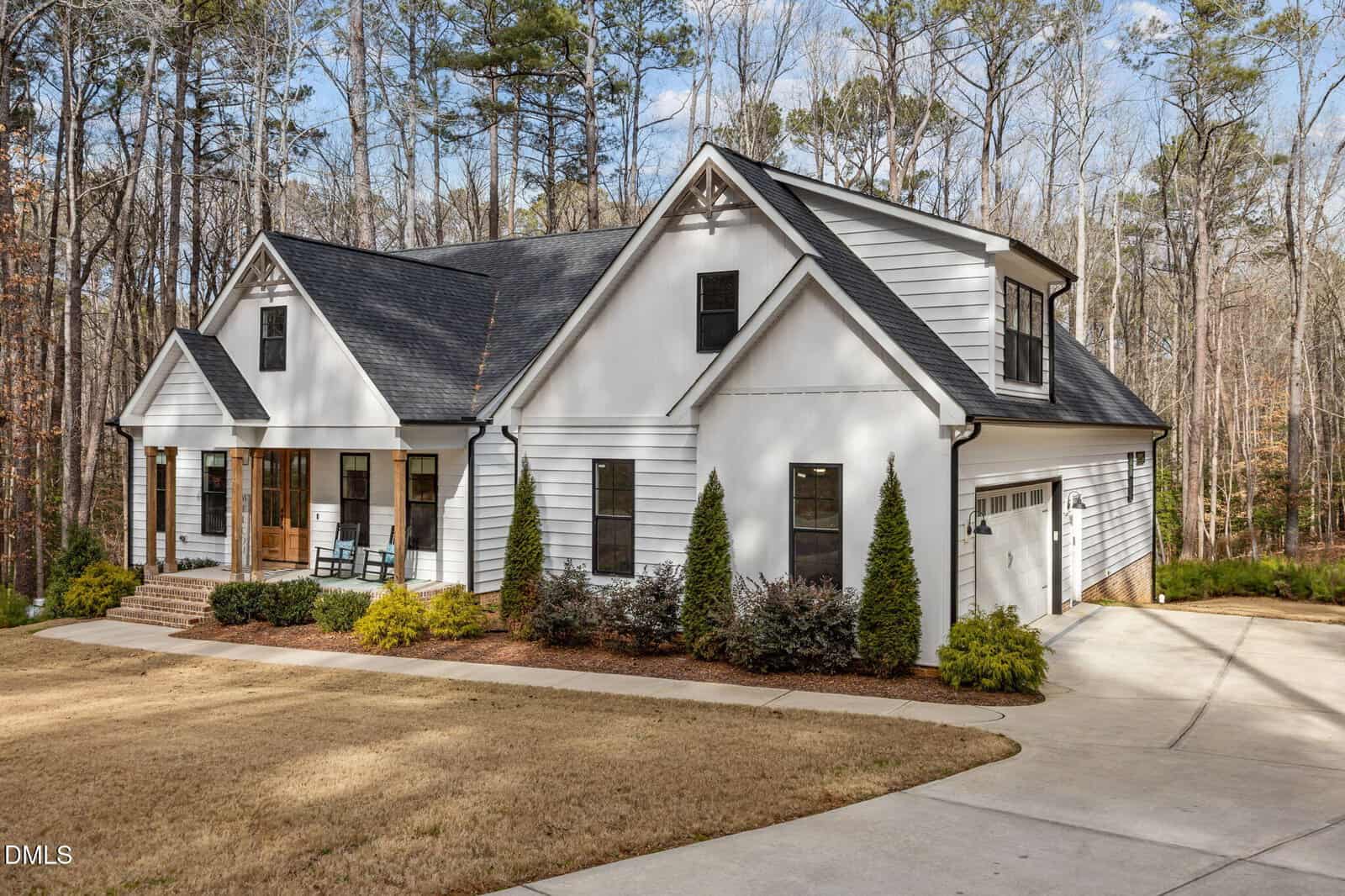 A modern white house with black trim, a gabled roof, front porch with rocking chairs, and a two-car garage, surrounded by trees and a neatly landscaped yard with bushes and a concrete driveway.