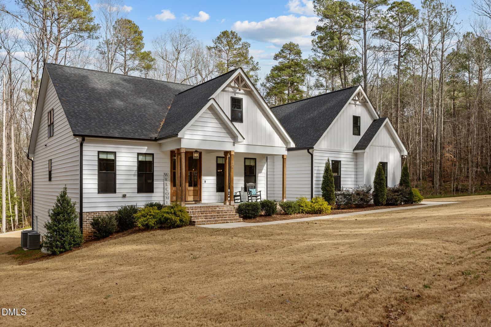 A modern white house with black trim, a covered front porch, and gabled roof sits on a large, grassy yard bordered by trees under a partly cloudy sky. Shrubs line the front of the house.