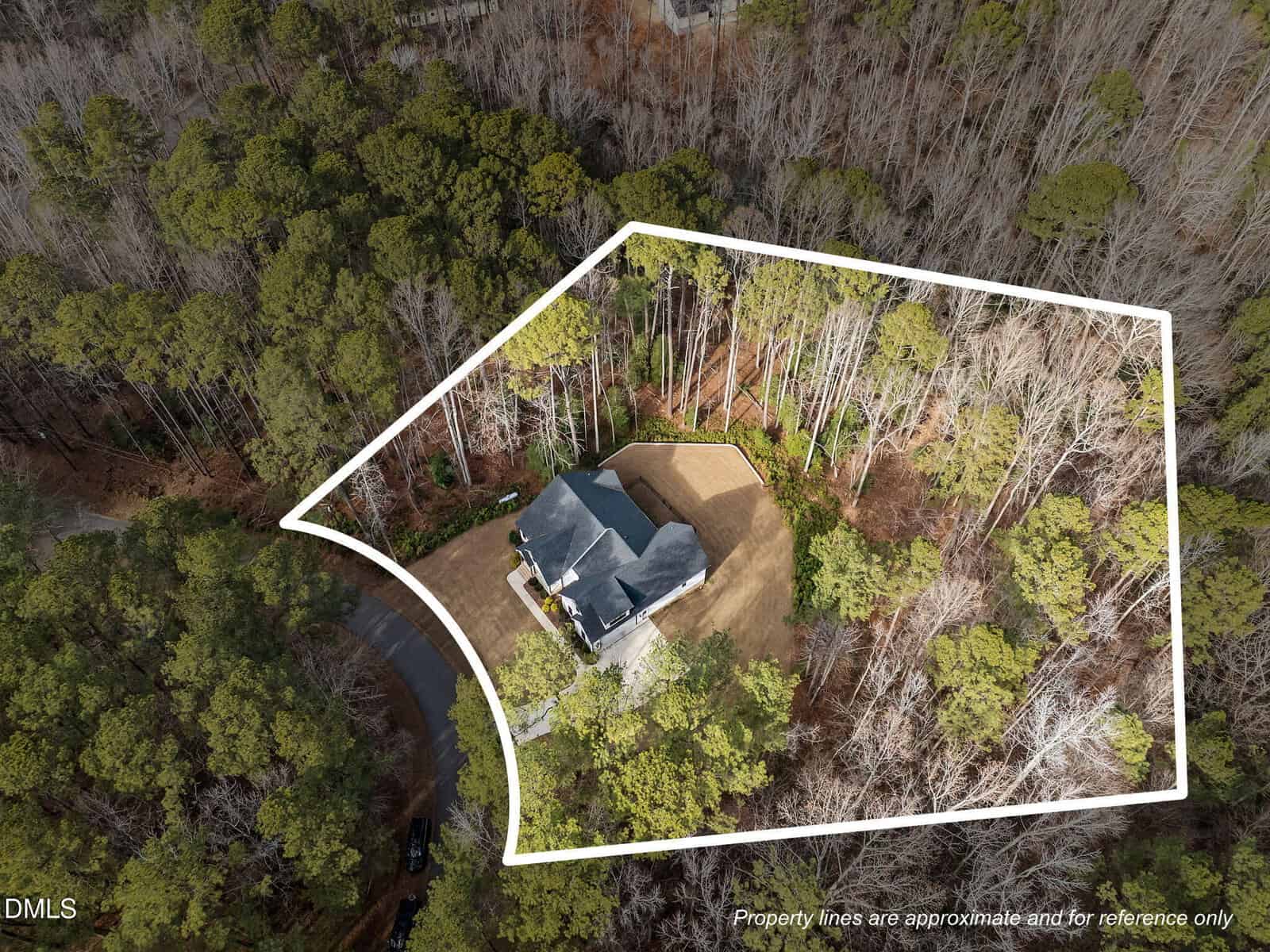 Aerial view of a house surrounded by trees, with a white outline marking the approximate property boundaries. The property sits at the edge of a forested area beside a curved road.