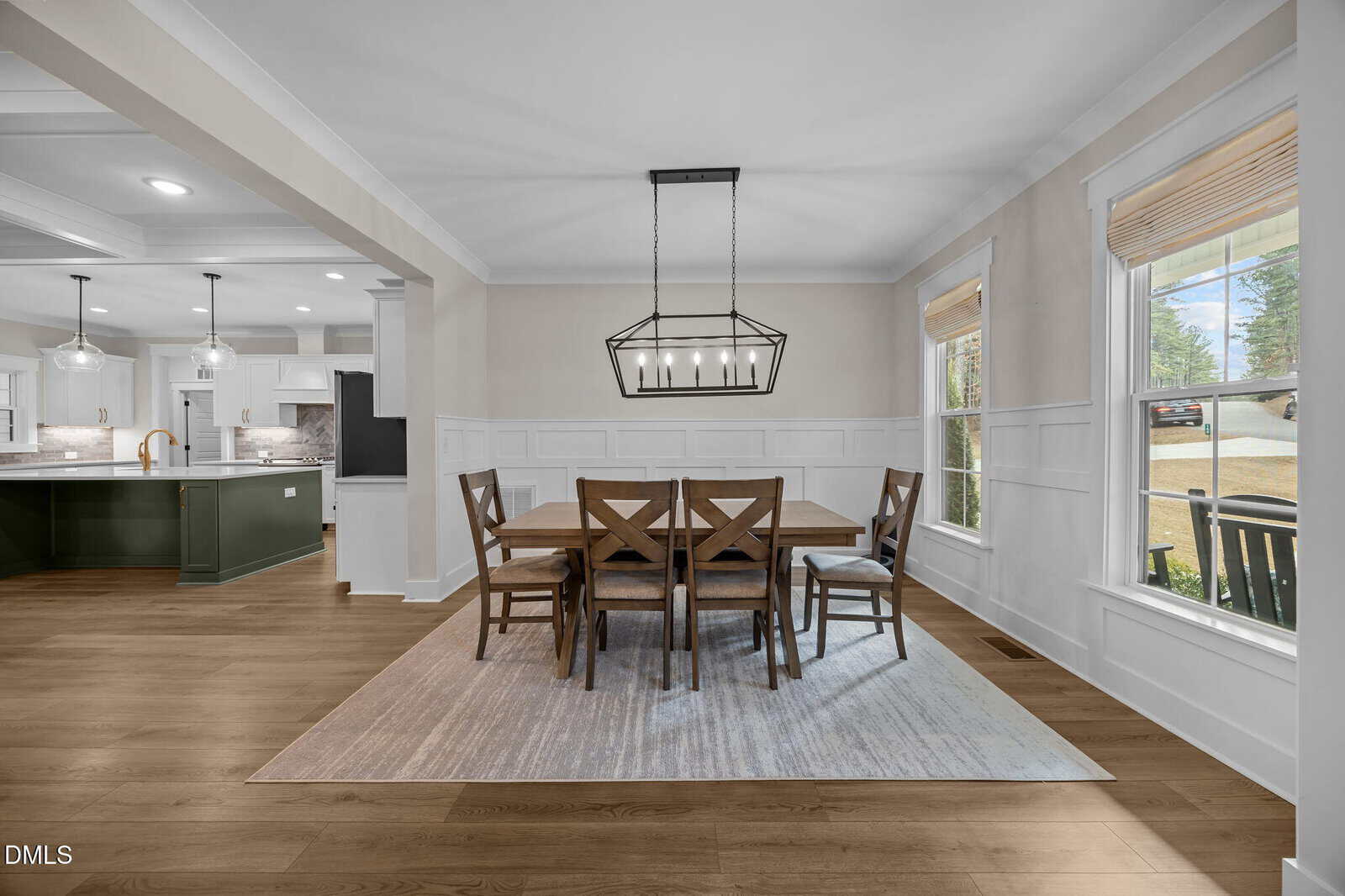 A dining room with a wooden table and six chairs on a rug, next to large windows. The adjacent kitchen features green cabinets, pendant lights, and a central island.