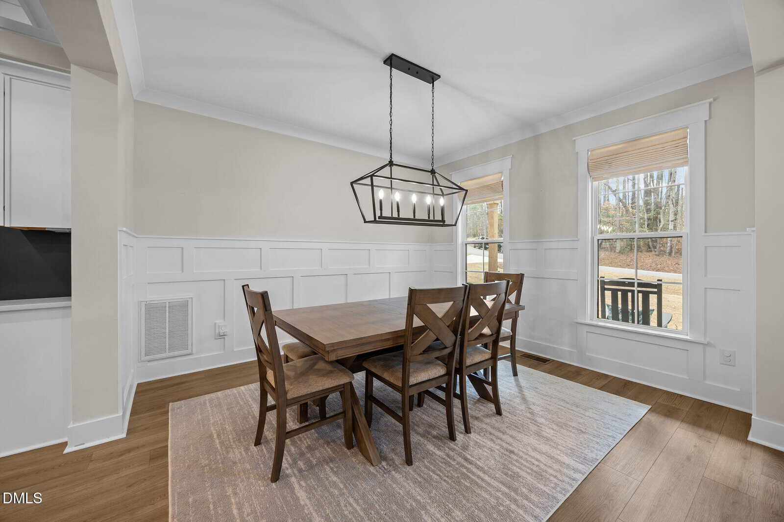 Modern dining room with a wooden table, six matching chairs, a geometric chandelier, large windows with natural light, white paneled walls, and hardwood floors. A neutral rug sits under the table.