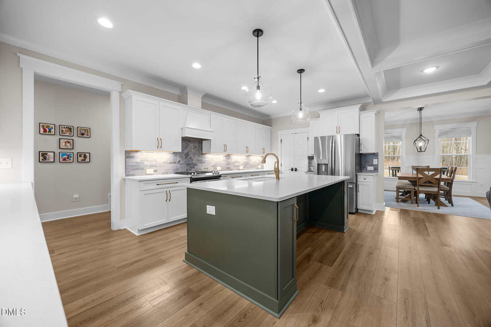 Modern kitchen with white cabinets, stainless steel appliances, a large green island with a white countertop, wood flooring, pendant lights, and an adjacent dining area with large windows.