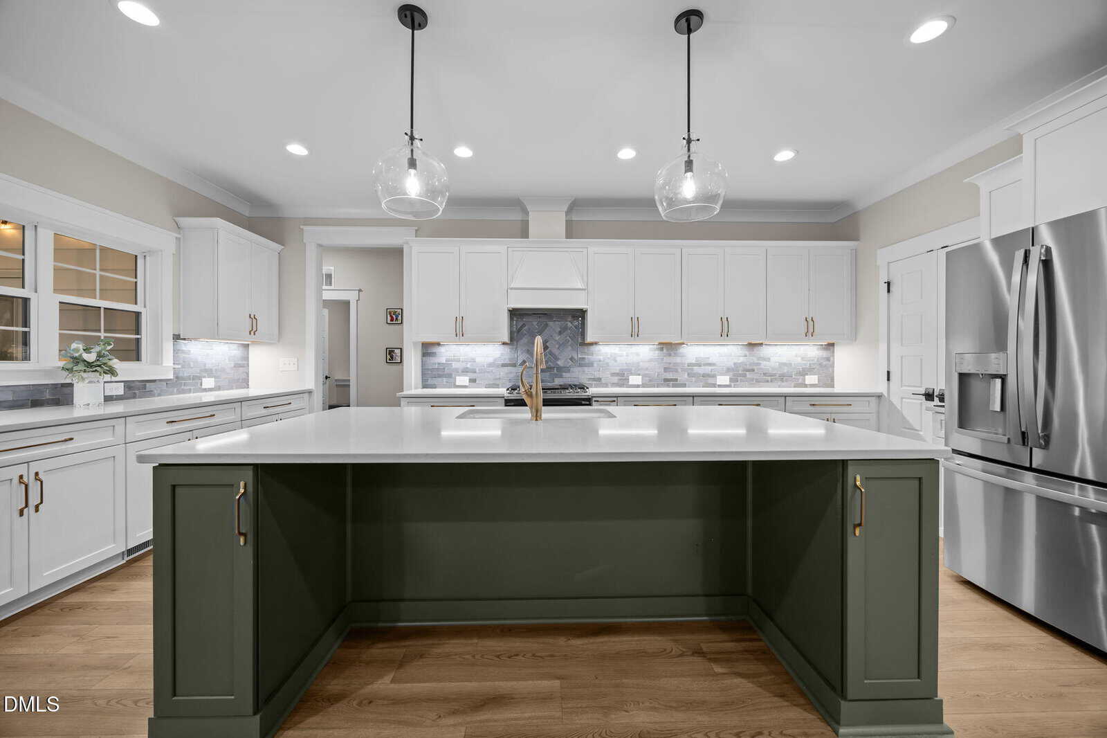 Modern kitchen with white cabinets, stainless steel appliances, a large green island with white countertop, glass pendant lights, wood flooring, and gray tile backsplash.