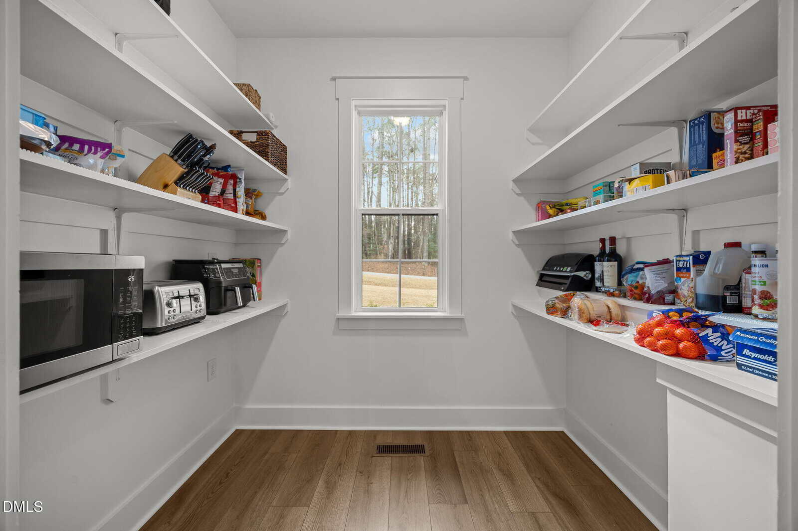 A bright pantry with white shelves holding appliances, canned goods, snacks, and produce. A window at the back lets in natural light, and the floor is wood.