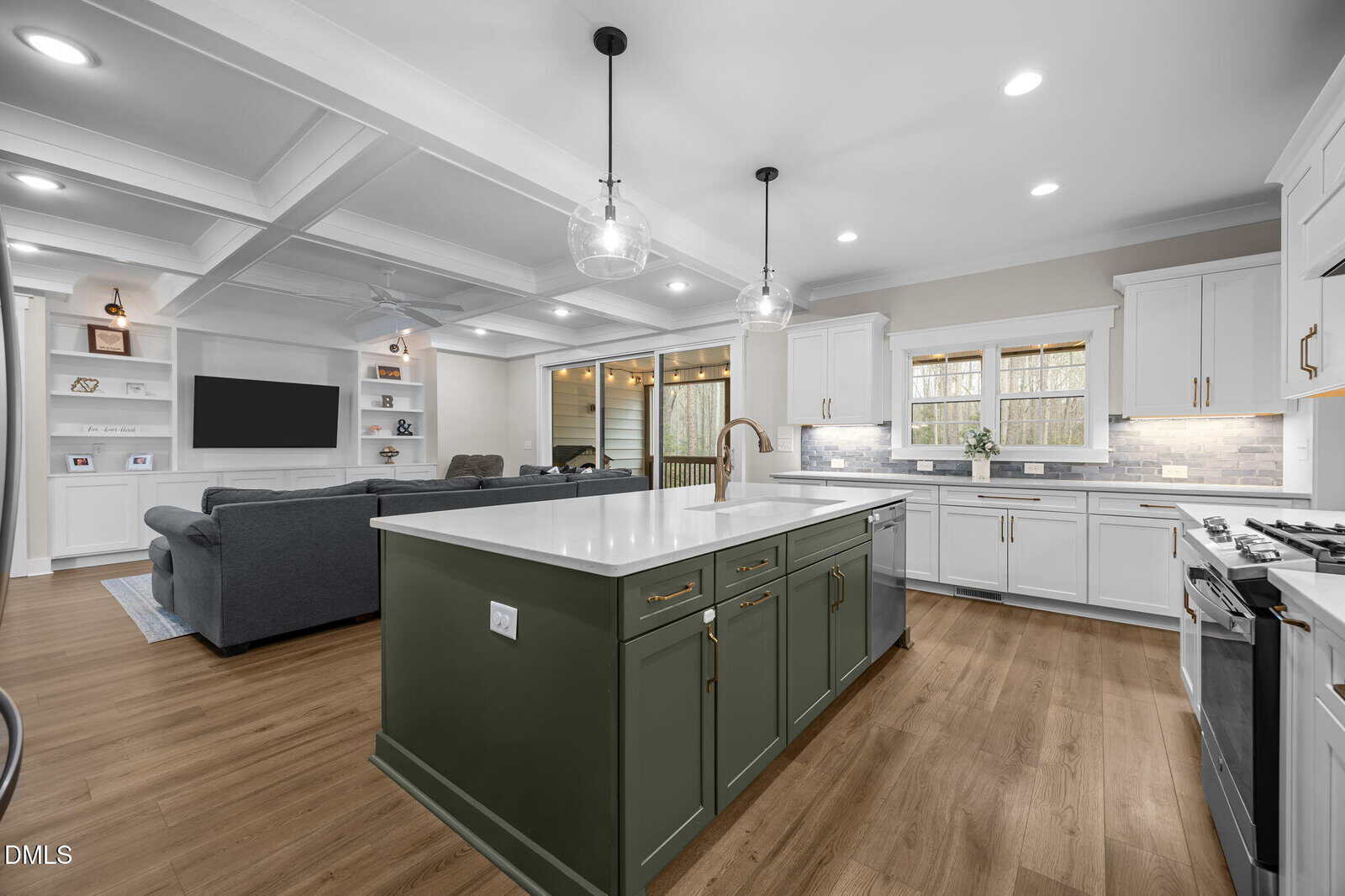 Modern kitchen with a large green island, white cabinets, marble countertops, stainless steel appliances, and pendant lights. The space opens to a living area with built-in shelves, a TV, and a gray sectional sofa.