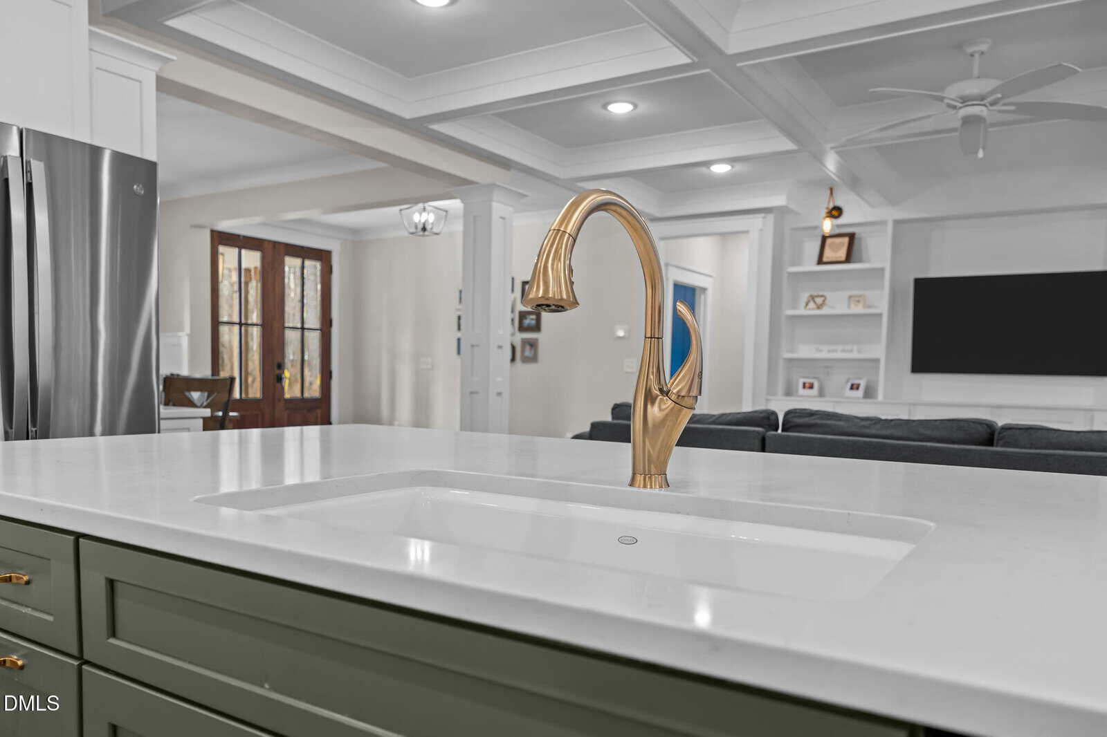 A modern kitchen with a gold faucet on a white island countertop, green cabinets with gold handles, and an open view into a living room with built-in shelves, a TV, and large double doors.