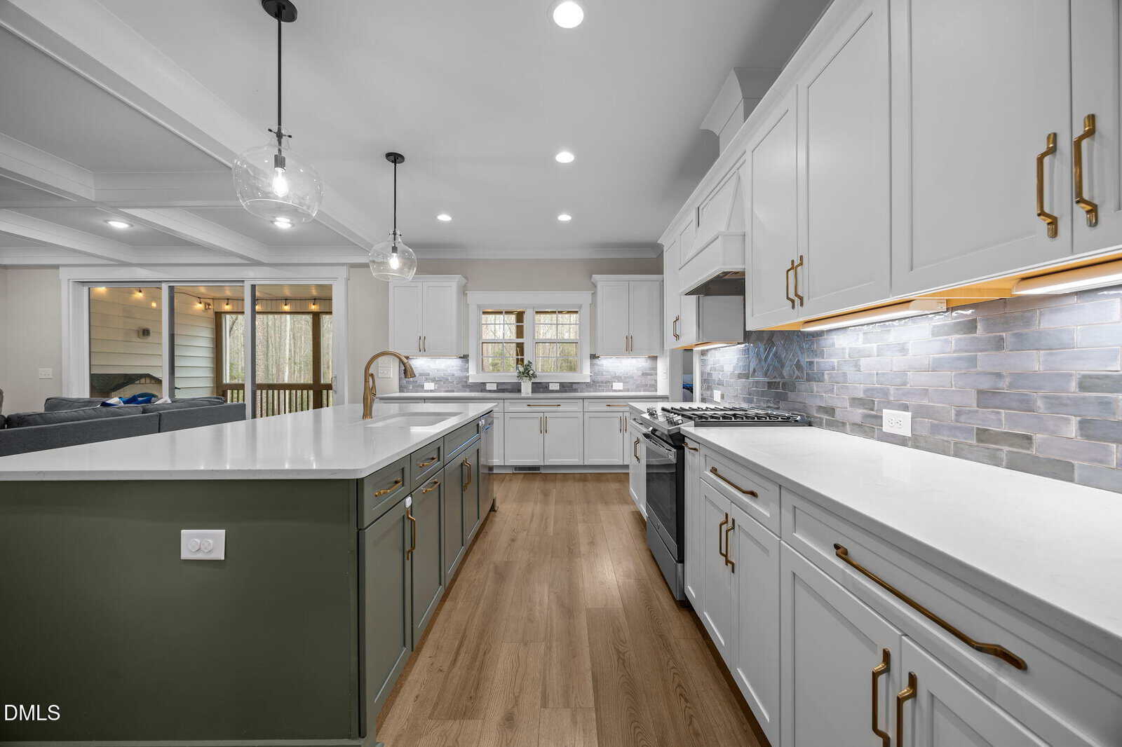 Modern kitchen with white cabinets, stainless steel appliances, green island with pendant lights, white countertops, wood floors, and a tile backsplash. Open layout connects to the living area and a deck through glass doors.