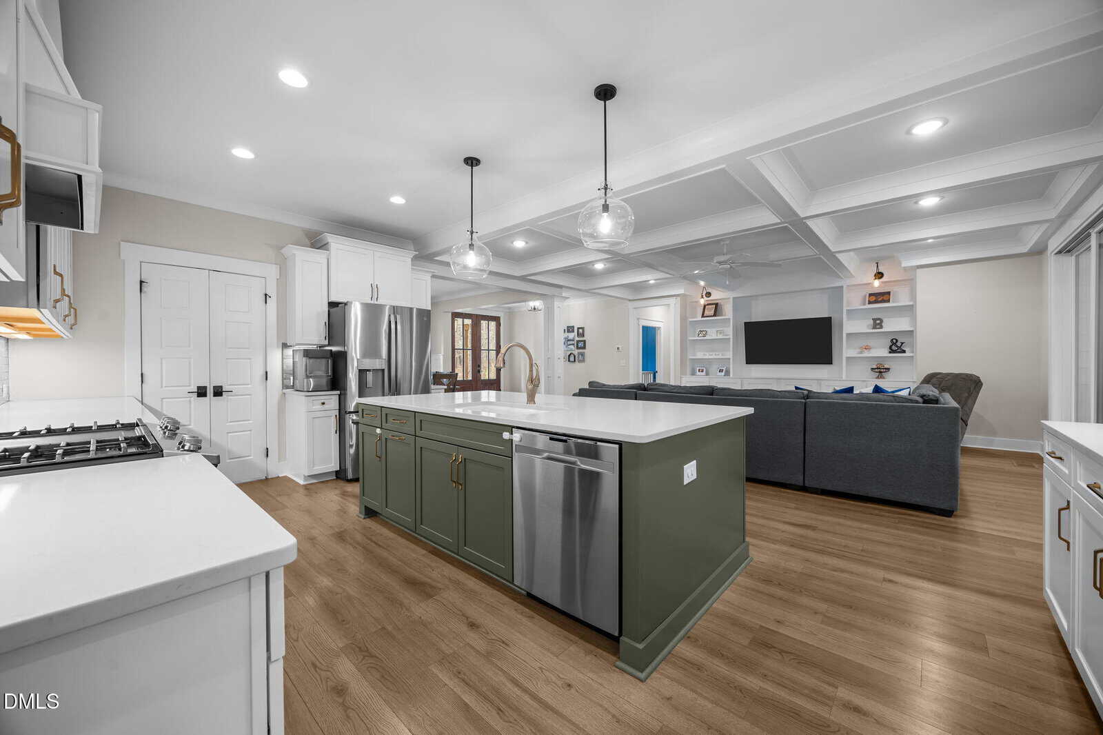 Modern open-concept kitchen and living room with wood floors, white cabinets, stainless steel appliances, a green island, pendant lights, and a large gray sectional facing a wall-mounted TV and built-in shelves.