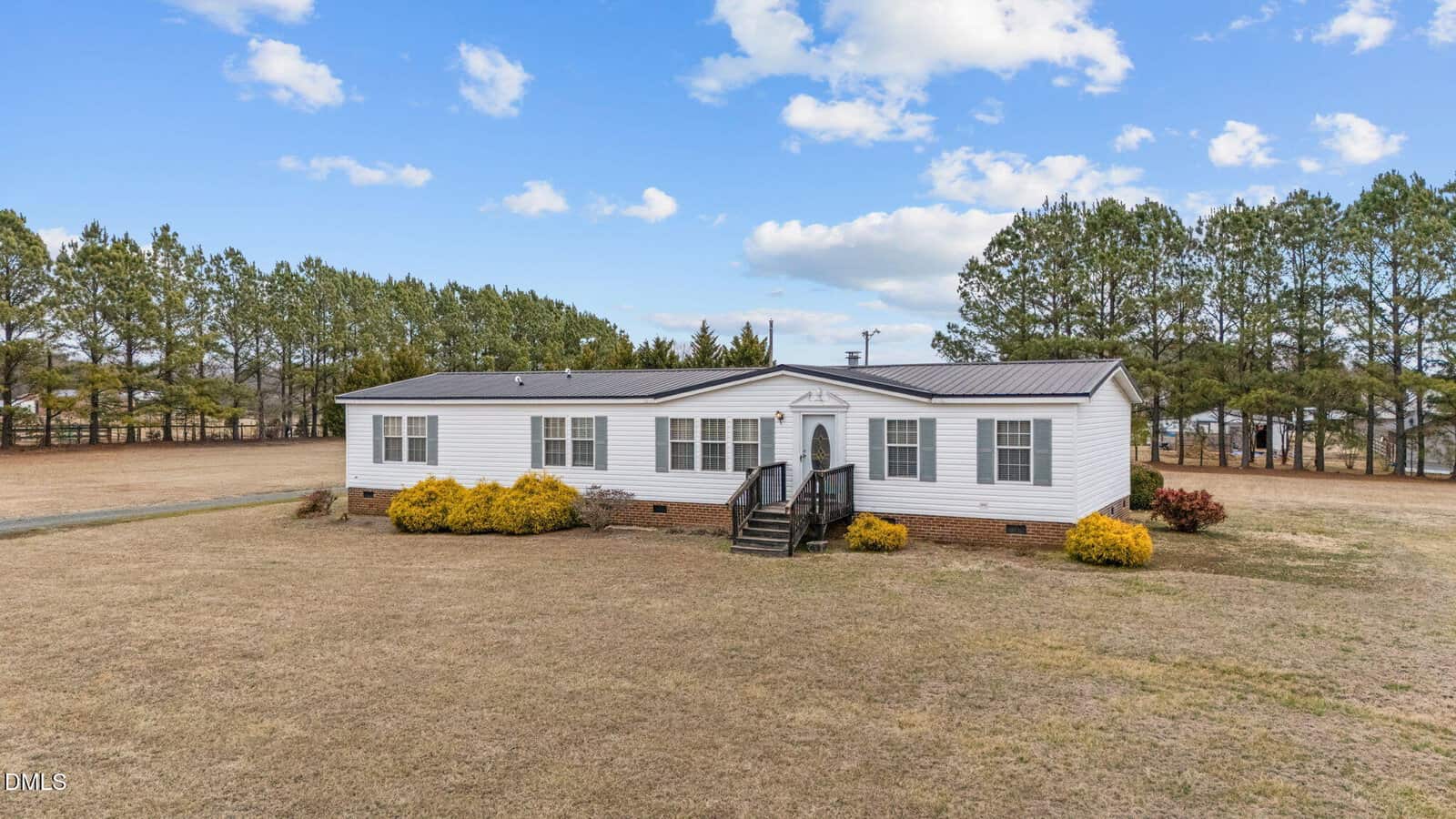 A white, single-story manufactured home with a black roof stands on a spacious, grassy lot. Shrubs border the front, and a row of tall pine trees lines the background under a partly cloudy sky.
