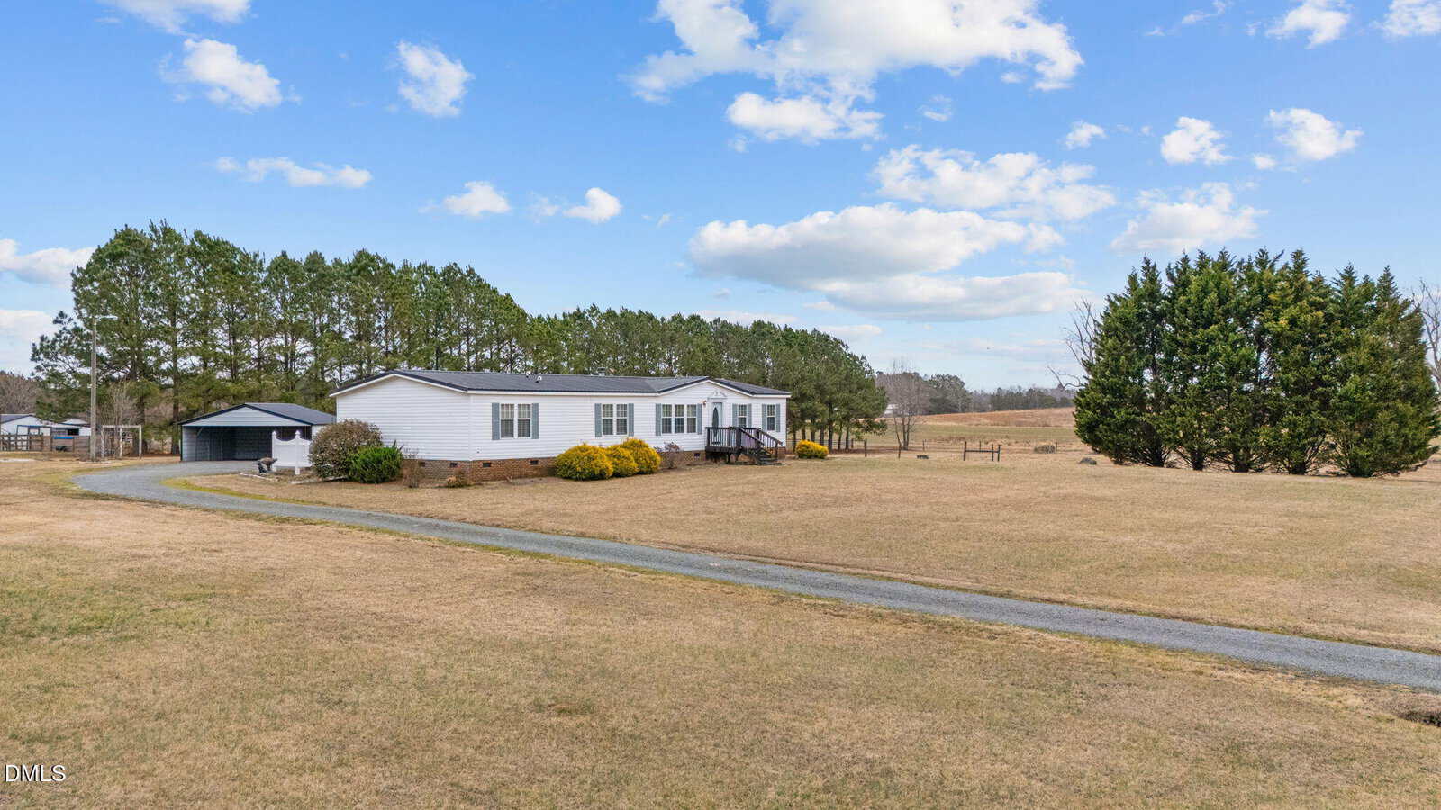 A white manufactured home sits on a large, open grassy lot with a gravel driveway, surrounded by trees under a blue sky with scattered clouds.